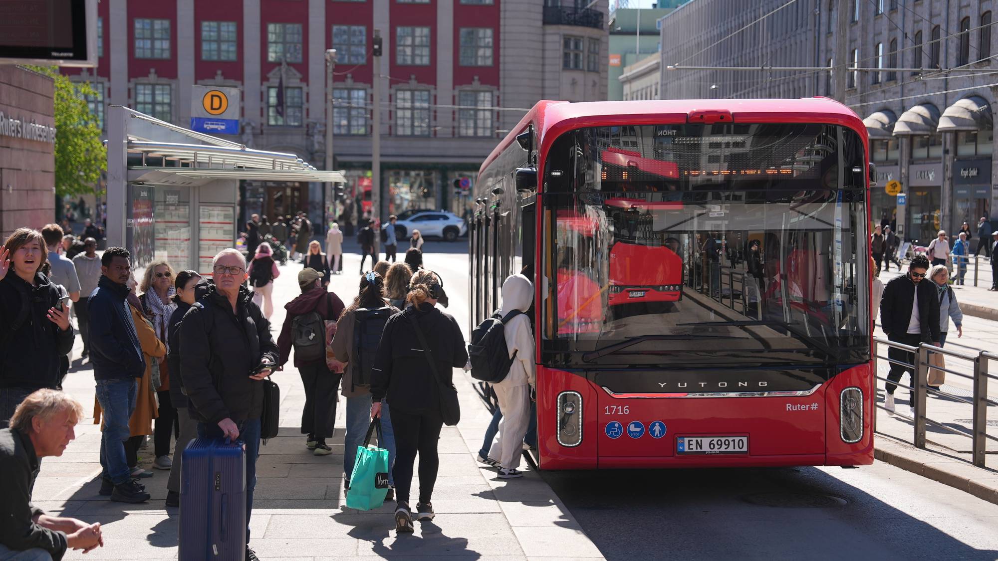 Folk venter på en rød buss. En buss med nummer 1716 og registreringsnummer EN 69910 står ved et busstopp. Passasjerer går av og på bussen. Omgivelsene er en travel bygate med bygninger og andre folk. (Bildebeskrivelsen er laget av en KI-tjeneste)