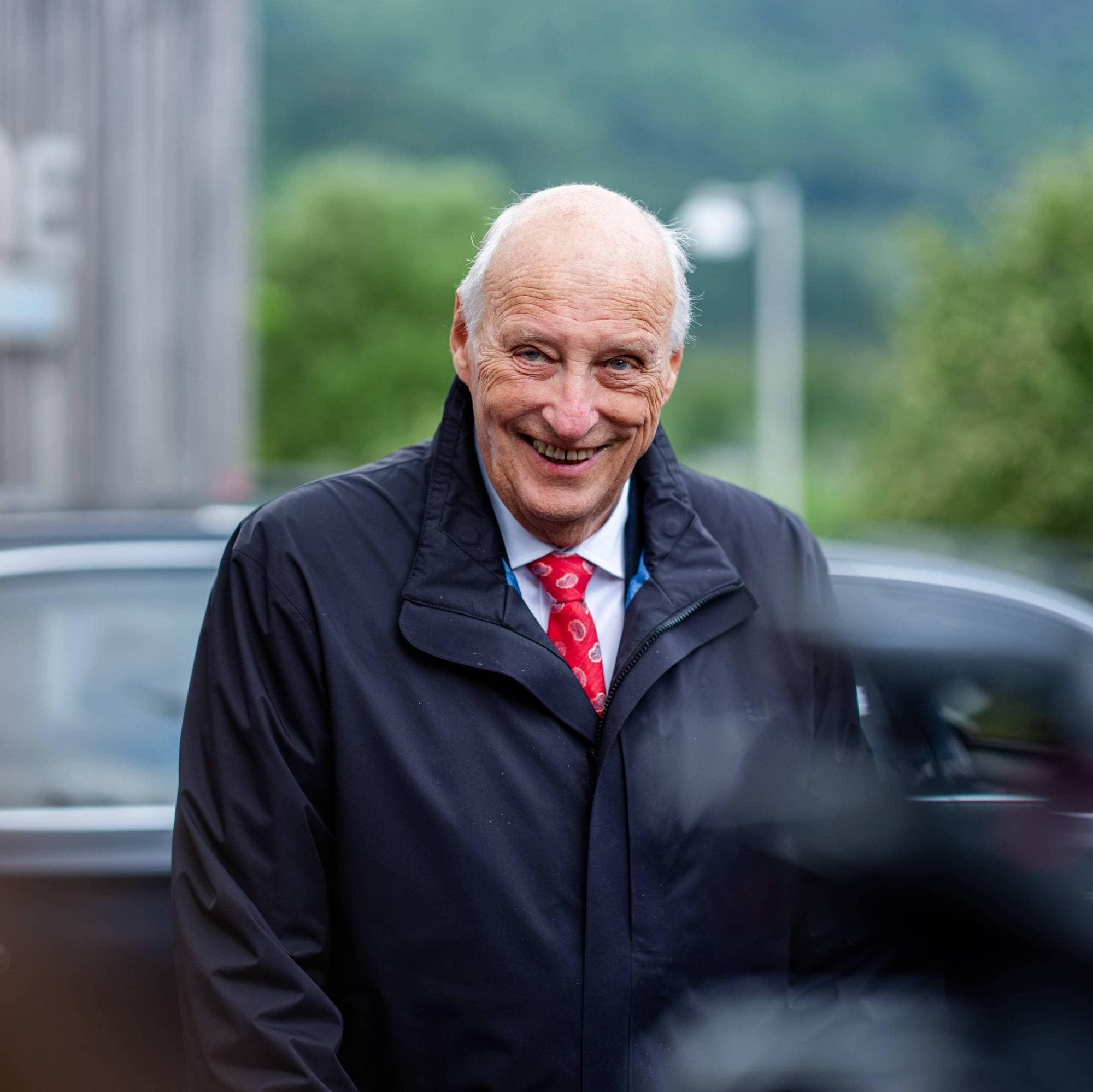 Kong Harald smiler mens han står utenfor Minne kultursenter i Åseral under kongeparets fylkesreise i Agder og Rogaland i mai 2024.