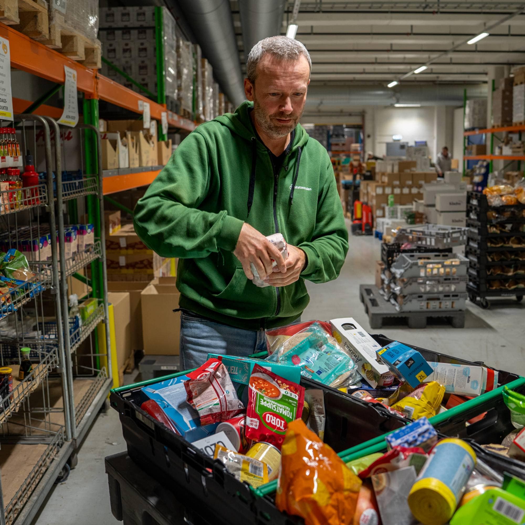 En mann i grønn hettegenser pakker matvarer i en handlekurv på et lager.