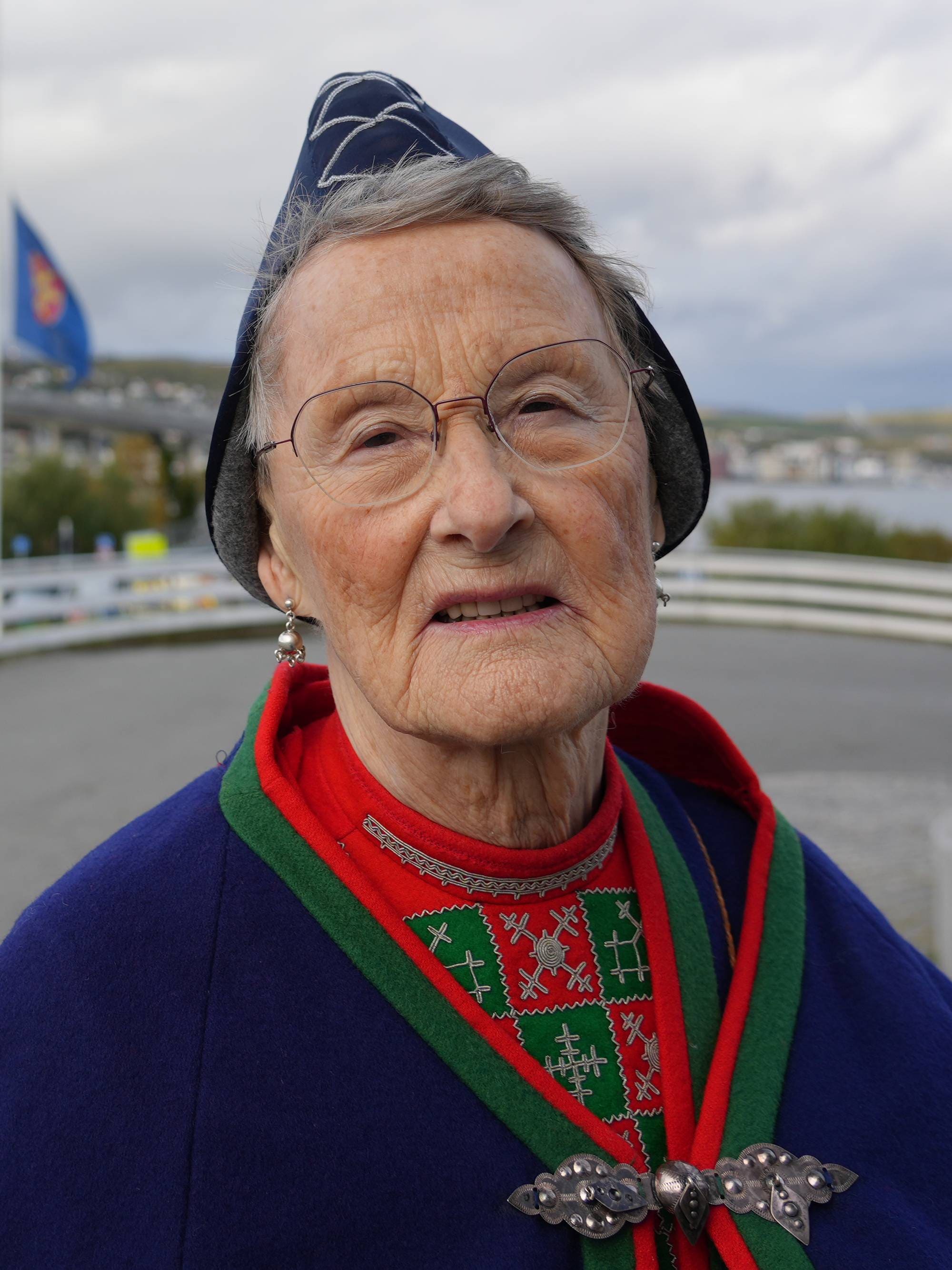 Eldre kvinne ikledd sørsamisk kofte i forgrunnen, med norsk, samisk og kvensk flagg heist på halv stang utenfor Tromsdalen kirke.