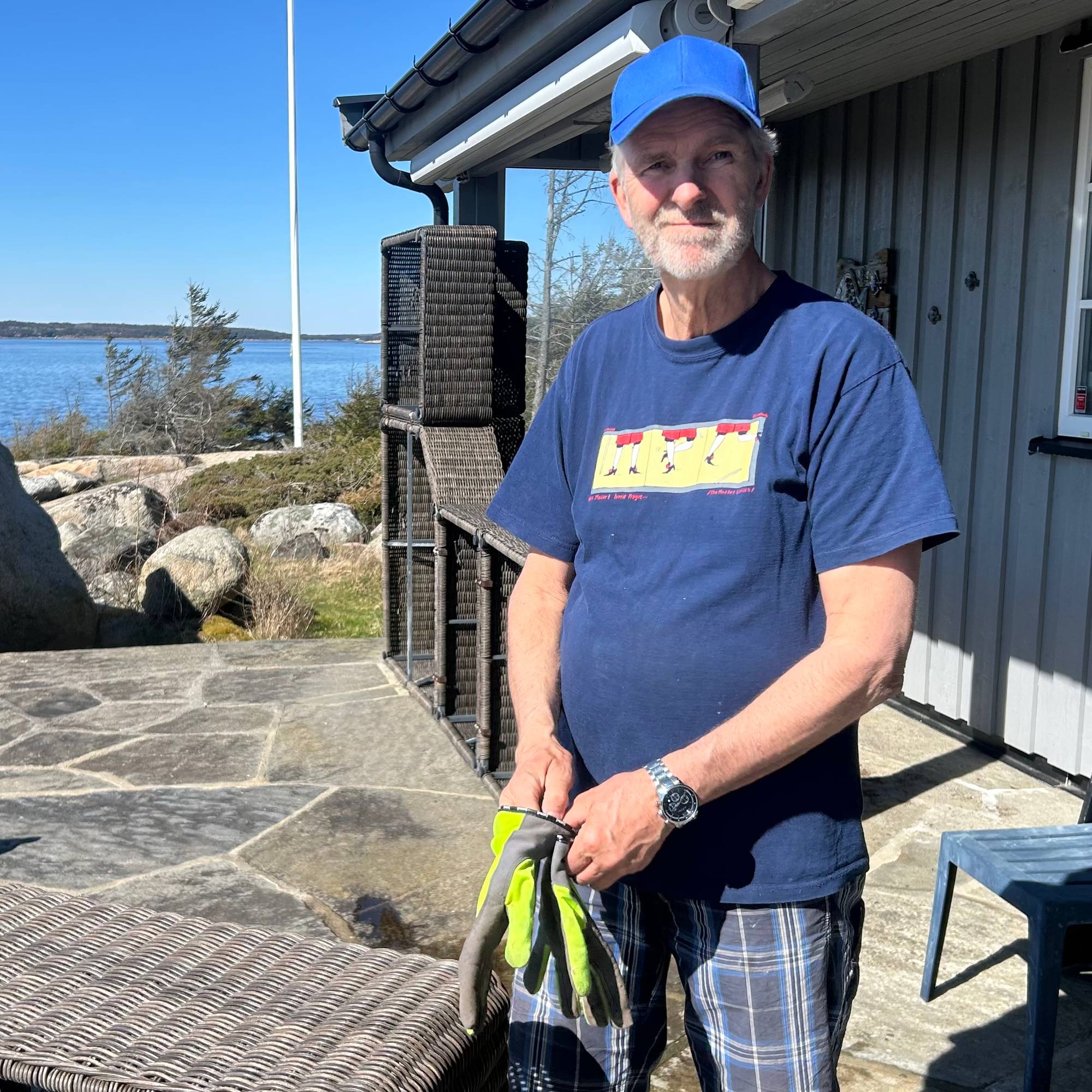 En mann med blå caps og mørkeblå t-skjorte står på en steinsatt uteplass. Han holder et par hansker i hendene.