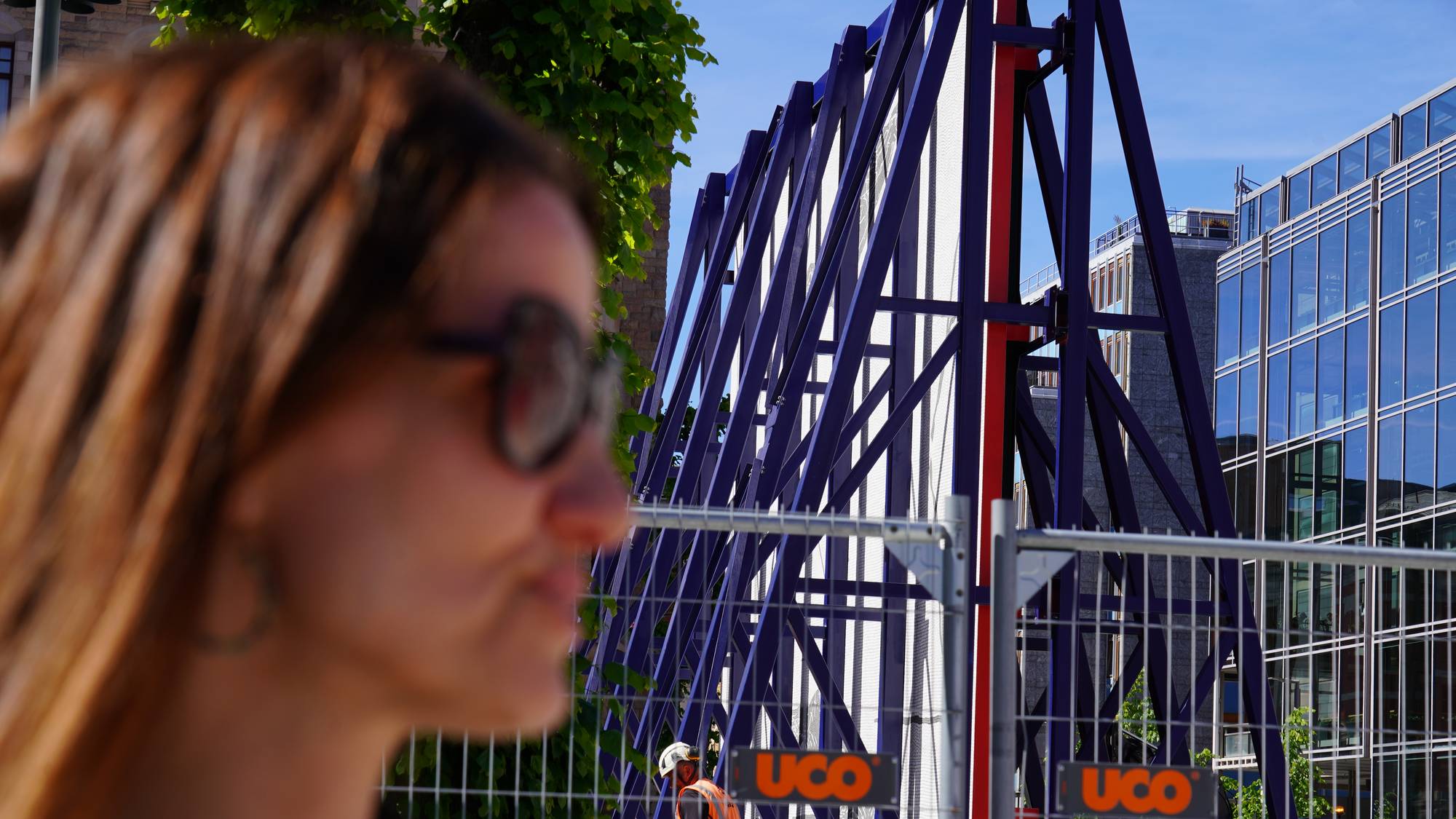 En kvinne er i forgrunnen, ute av fokus. Bak henne ser man en byggeplass. En arbeider i oransje verneutstyr er synlig. Det er et blått stillas bak et gjerde. (Bildebeskrivelsen er laget av en KI-tjeneste)