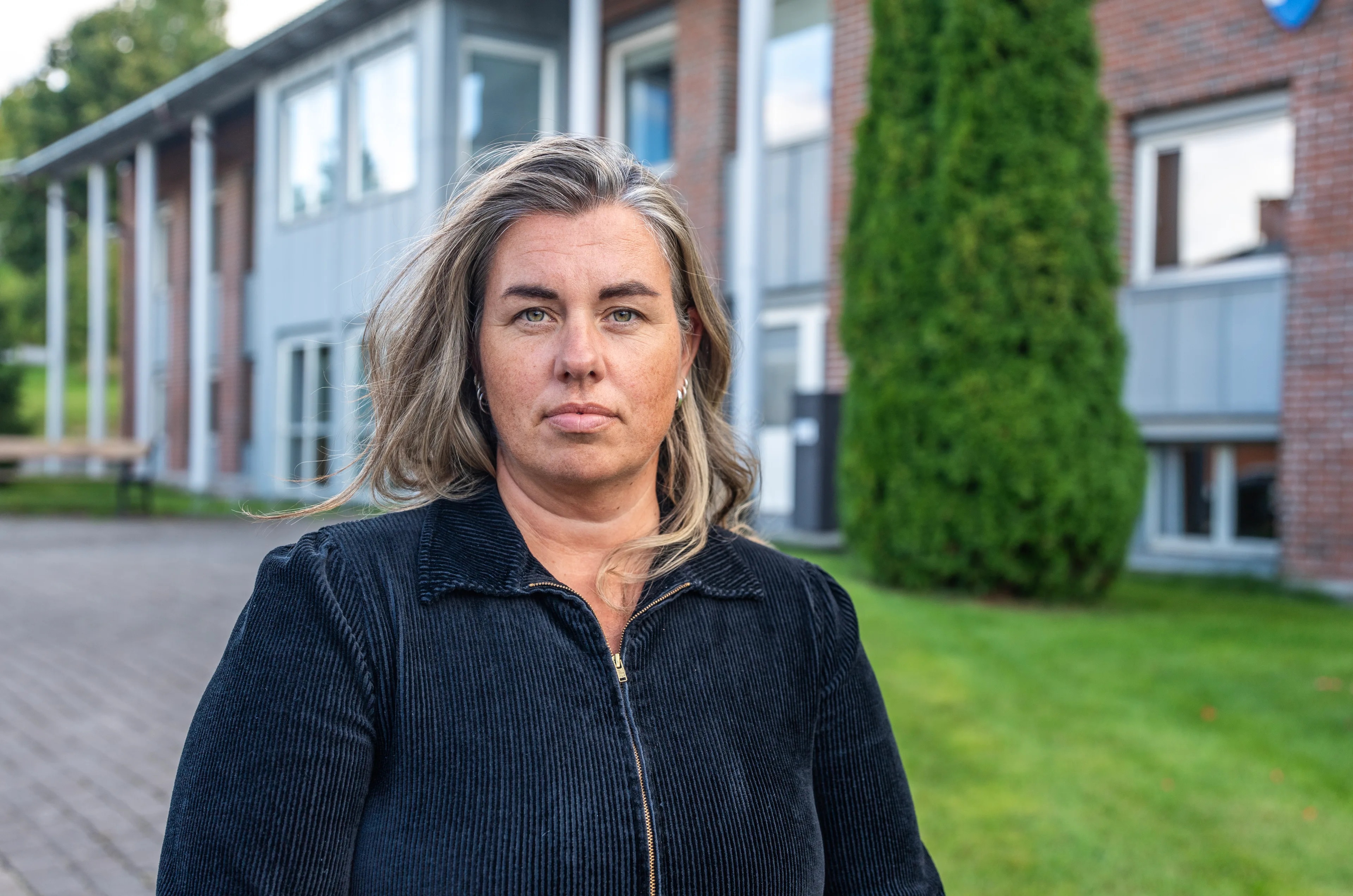 Kvinne står foran en teglsteinsbygning. Hun ser mot kamera med et nøytralt uttrykk. (Bildebeskrivelsen er laget av en KI-tjeneste)