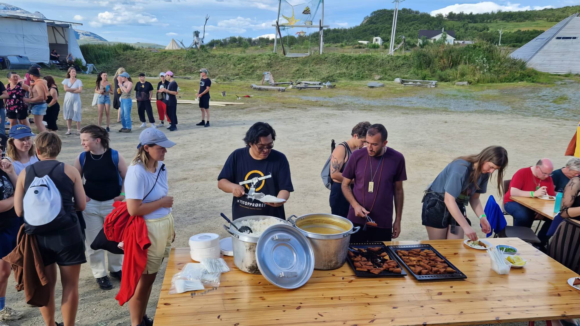 Folk står i kø ved et utendørs serveringsbord med mat under en festival. (Bildebeskrivelsen er laget av en KI-tjeneste)