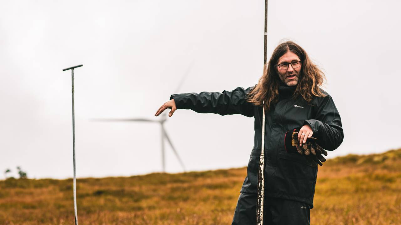 Forsker Anders Lyngstad står på en myr i Roan vindkraftverk. Han peker og forklarer, mens han holder et bor som brukes for å ta torvprøver. I bakgrunnen skimtes en vindturbin.