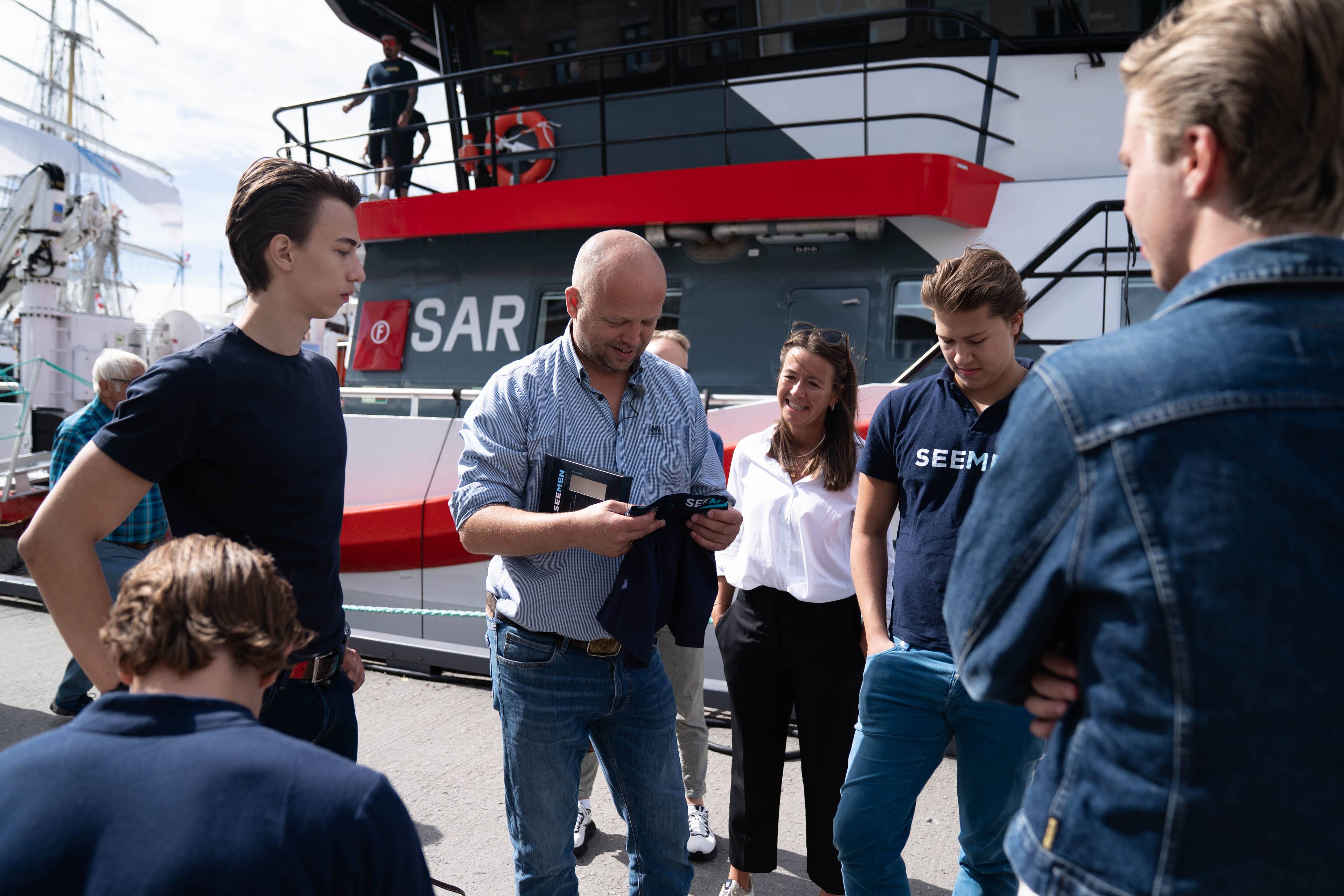 Kronprins Haakon viser frem en t-skjorte til en gruppe ungdommer foran en redningsskøyte. (Bildebeskrivelsen er laget av en KI-tjeneste)