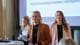 Caroline Bækkelund Hauge / NRK Tre kvinner står på en scene foran en prosjektor. Kvinnen i midten holder et hefte i hånden og ser mot publikum. Kvinnen til høyre har på seg en rosa blazer. Det vises en lys projeksjonsflate i bakgrunnen med tekst. (Bildebeskrivelsen er laget av en KI-tjeneste)
