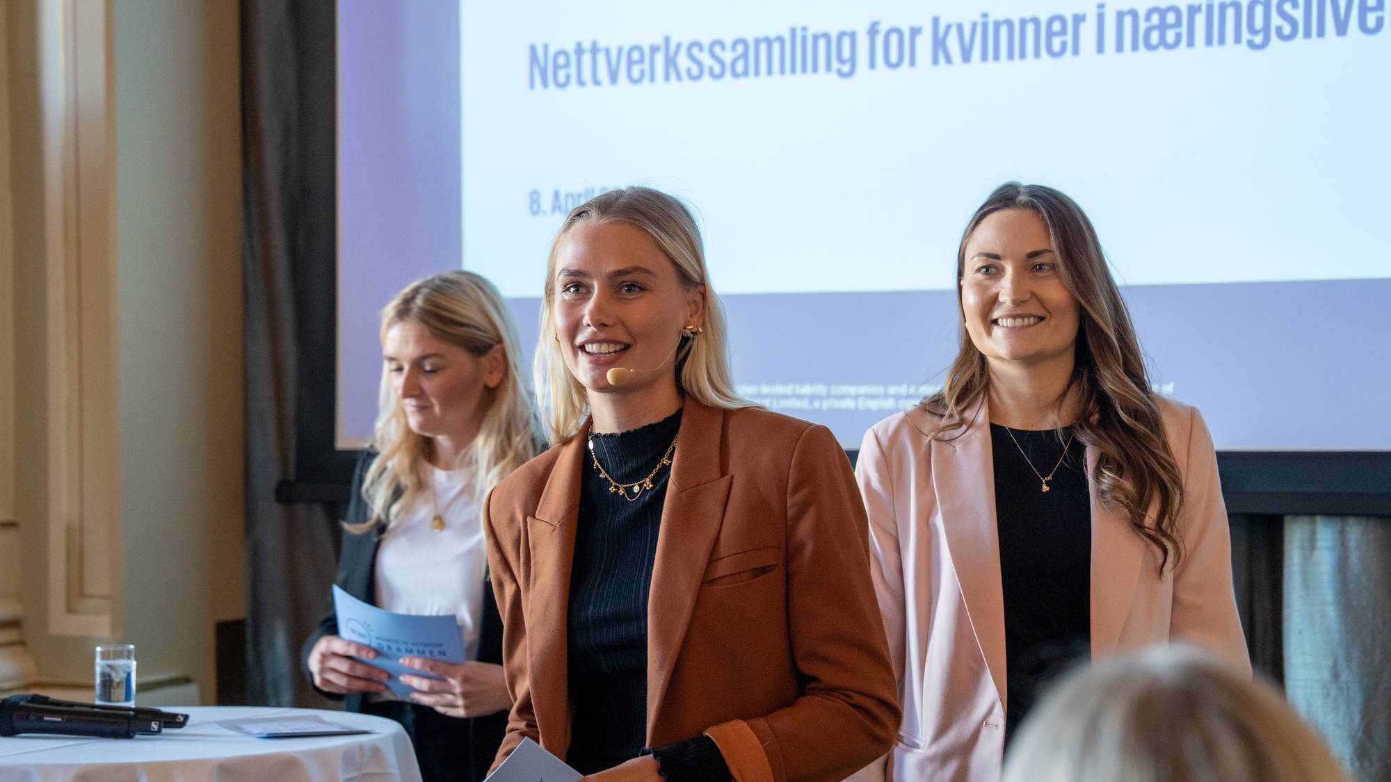 Tre kvinner står på en scene foran en prosjektor. Kvinnen i midten holder et hefte i hånden og ser mot publikum. Kvinnen til høyre har på seg en rosa blazer. Det vises en lys projeksjonsflate i bakgrunnen med tekst. (Bildebeskrivelsen er laget av en KI-tjeneste)