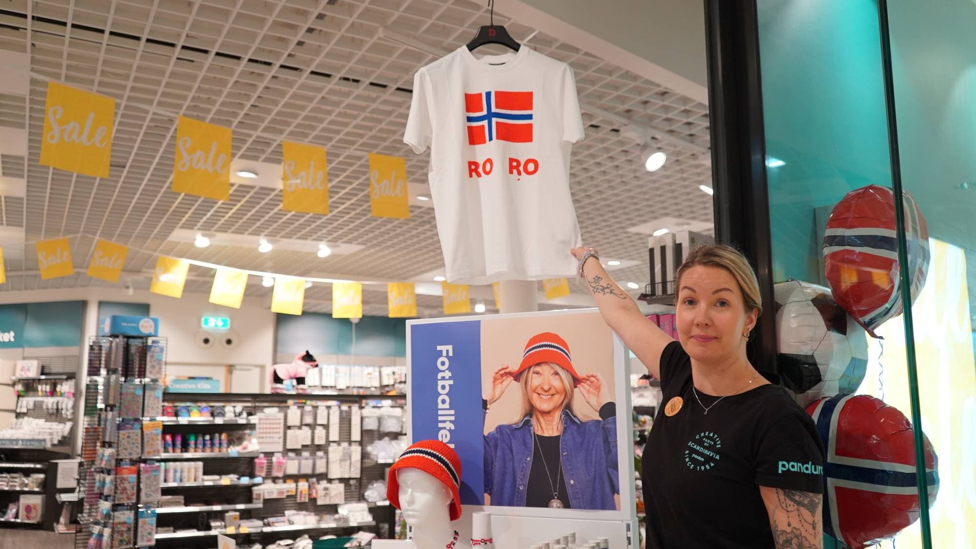 Butikkansatt holder opp en hvit t-skjorte med norsk flagg og teksten RO RO i en Panduro-butikk. 