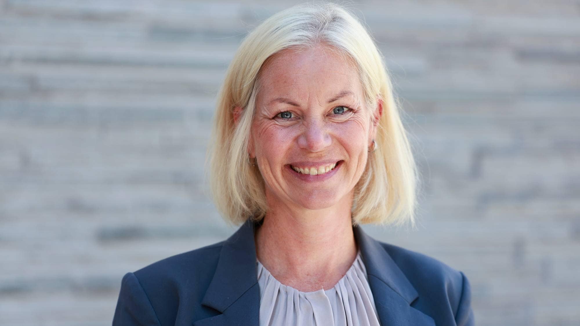 Portrett av en smilende kvinne med blondt hår i en mørk dressjakke foran en grå vegg. (Bildebeskrivelsen er laget av en KI-tjeneste)