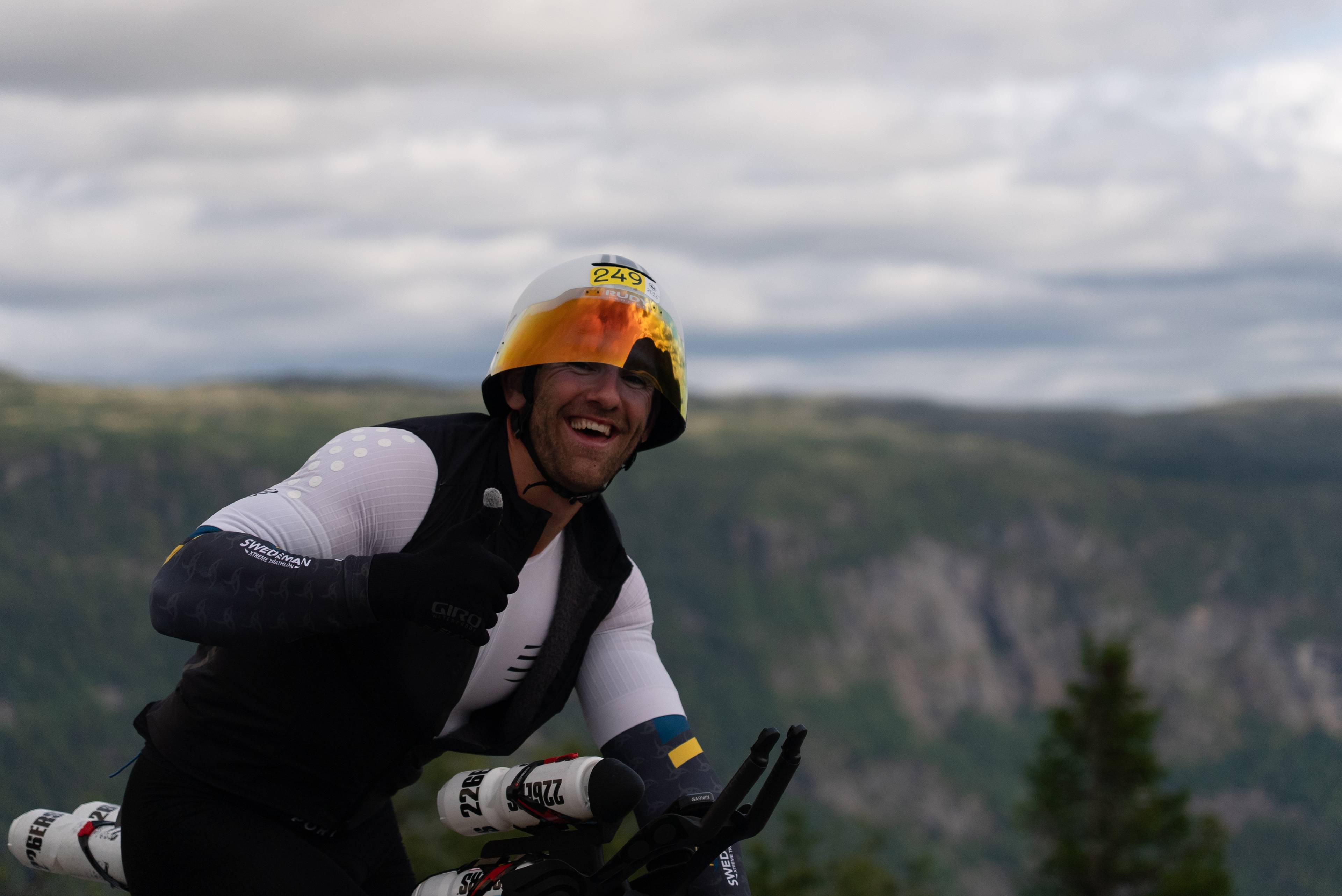 Syklist med hjelm og visir gir tommel opp i et fjellandskap. (Bildebeskrivelsen er laget av en KI-tjeneste)