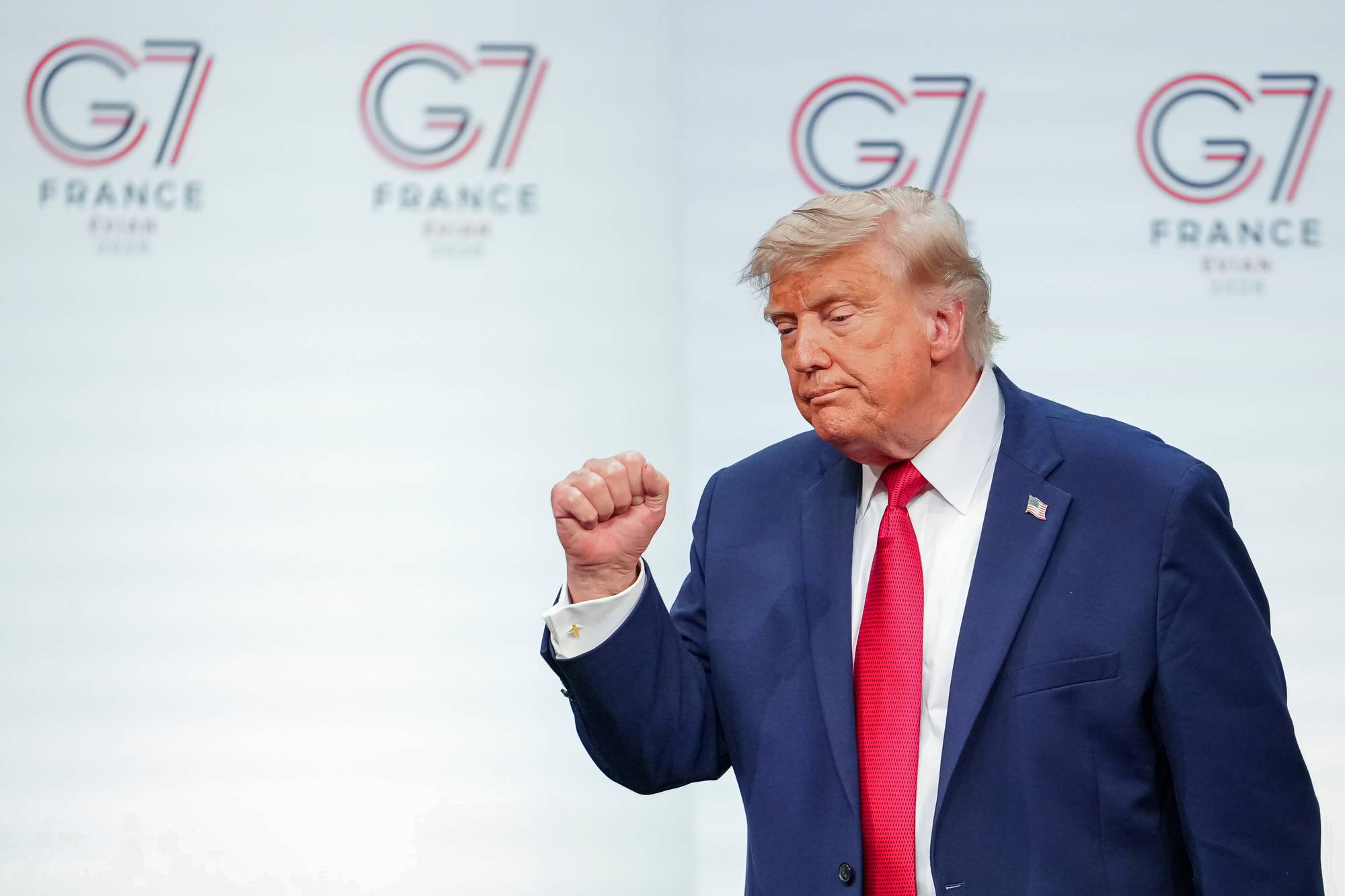 Donald Trump, kledd i mørkeblå dress og rødt slips, entrer en scene foran en bakgrunn med G7-logo. Han løfter den venstre knyttneven. Bakgrunnen viser flere G7-logoer og teksten "FRANCE 2019".