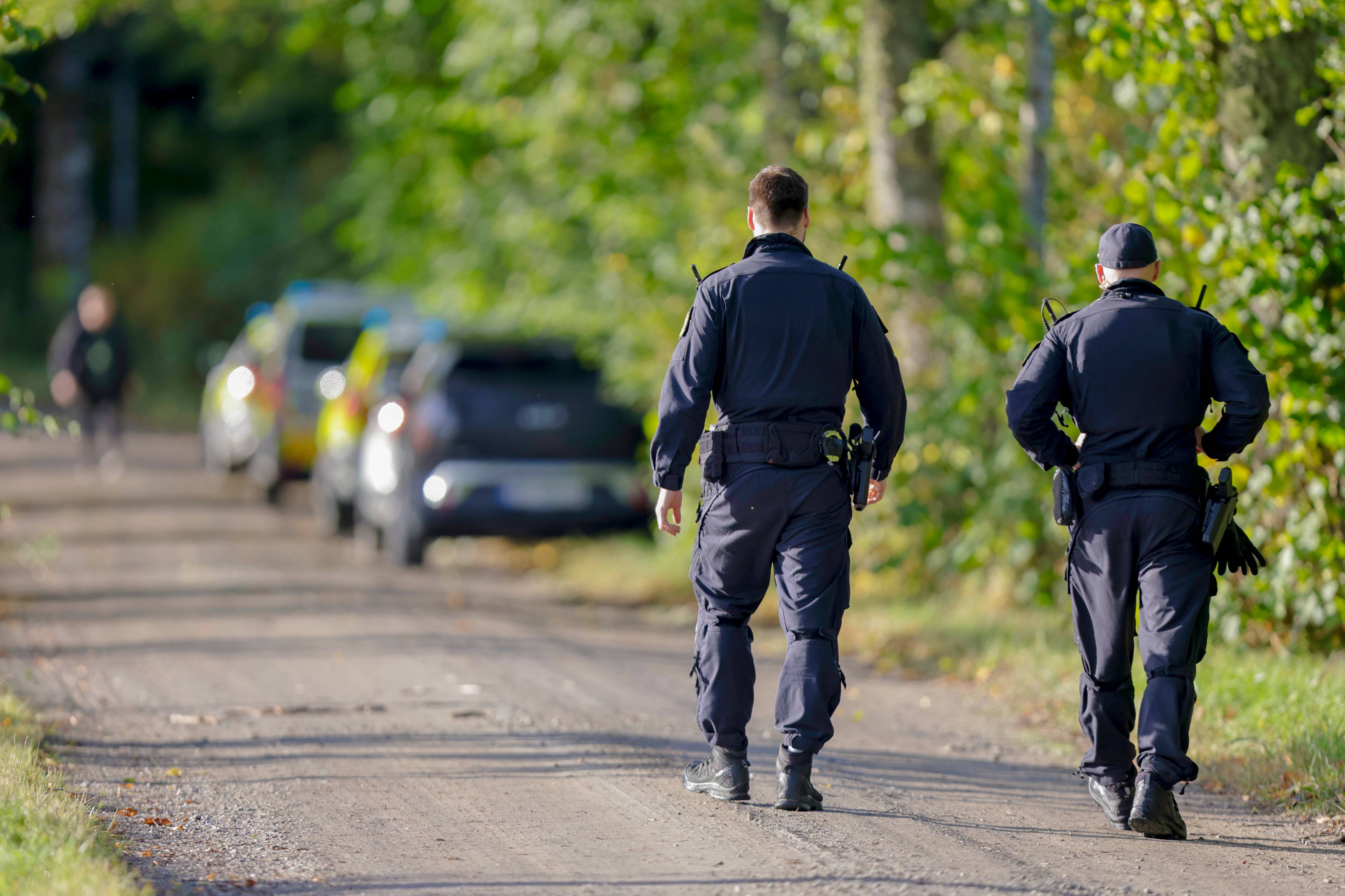 To politibetjenter går bortover en grusvei med politibiler i bakgrunnen. (Bildebeskrivelsen er laget av en KI-tjeneste)