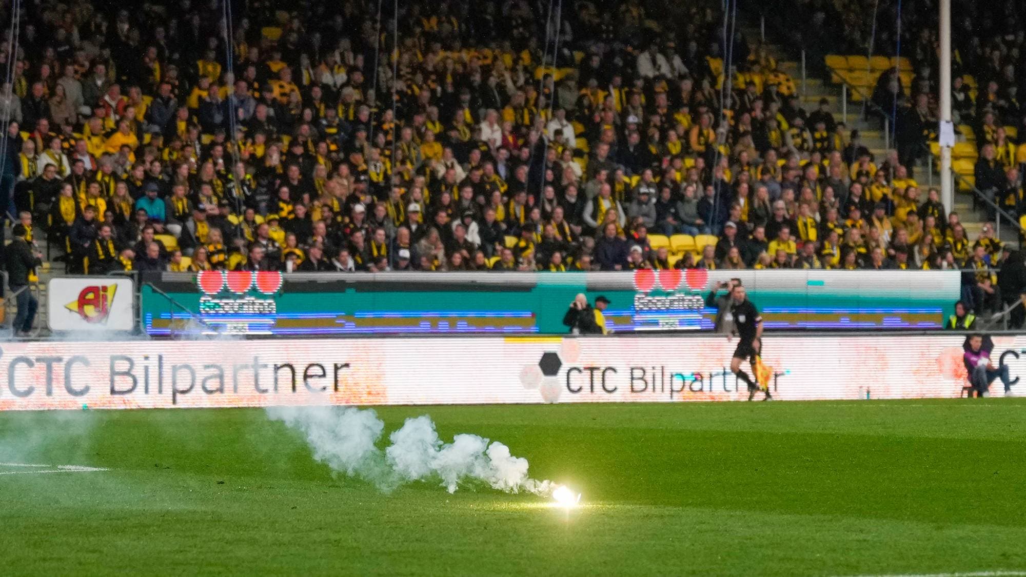 Fotballkeeper står på banen foran tribune med publikum mens et bluss brenner på gresset. (Bildebeskrivelsen er laget av en KI-tjeneste)