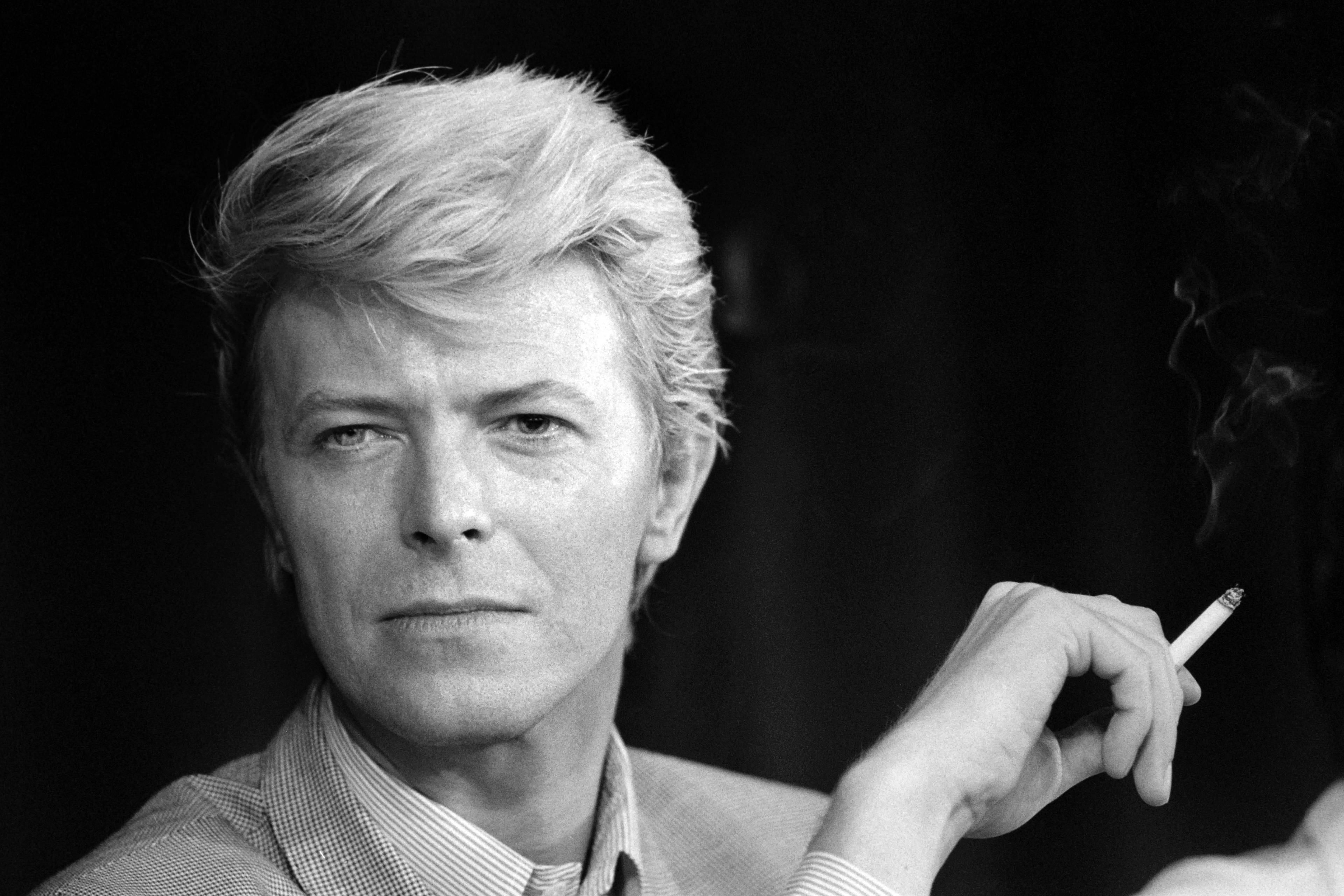 David Bowie i ein svart-kvitt fotografering. Han har blondt hår og ser mot kamera. Han holder ein sigarett i høyre hand, som ryk. (Bildebeskrivelsen er laget av ein KI-teneste)