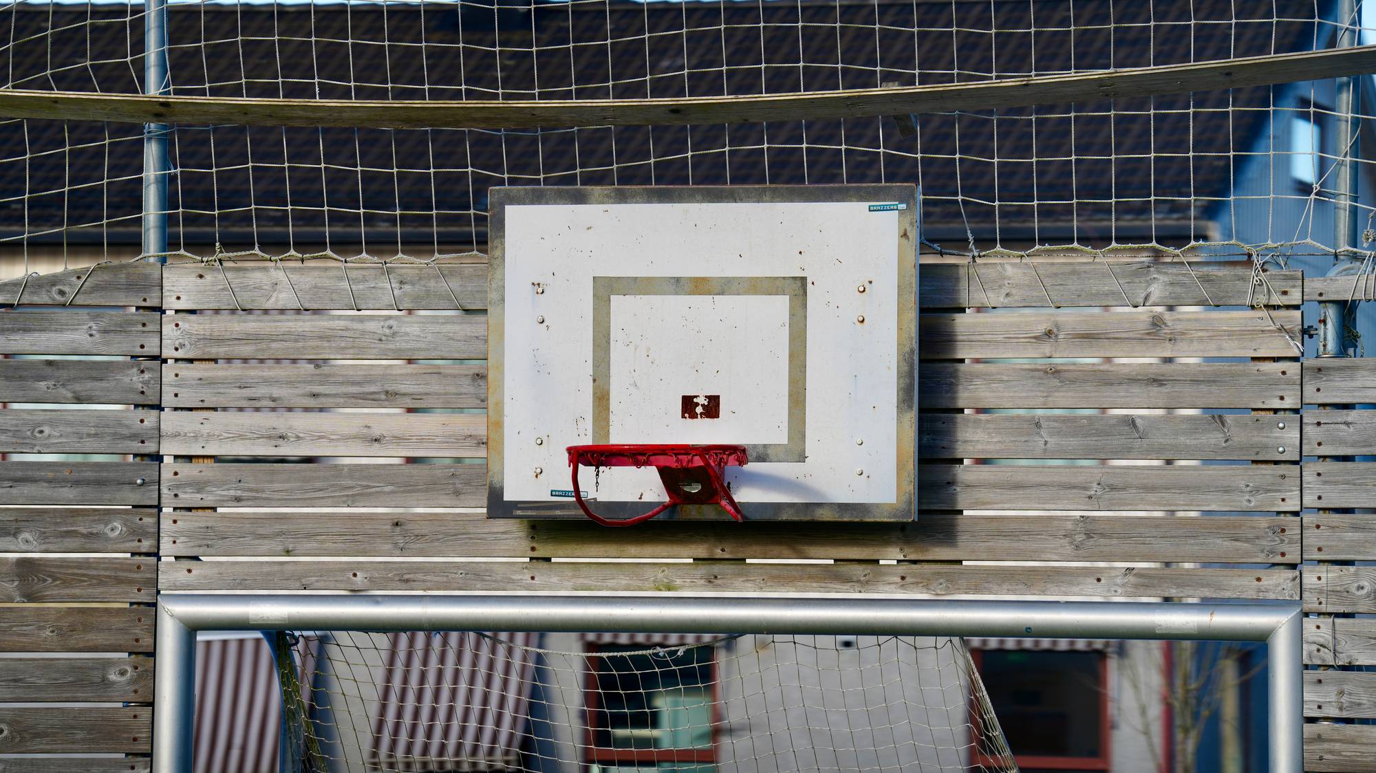 En basketballkurv med slitt hvit plate montert på en trevegg ved en ballbinge. (Bildebeskrivelsen er laget av en KI-tjeneste)