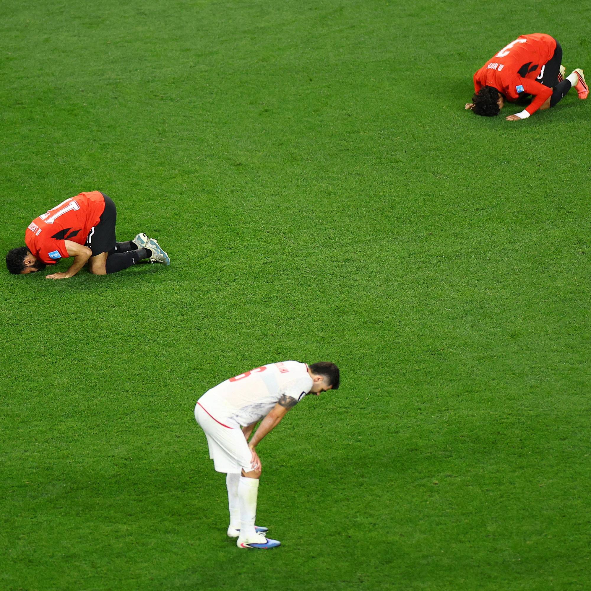 Tre menn på en grønn fotballbane. To av dem i oransje drakt har hodet i bakken.