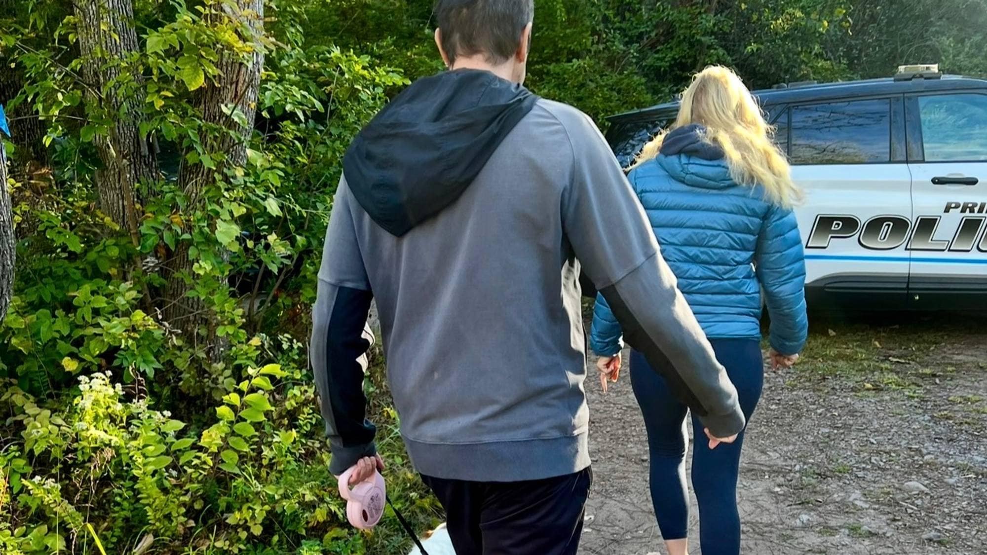 Et par og hunden deres går langs en grussti. En politibil er parkert i bakgrunnen. Personene er sett bakfra. (Bildebeskrivelsen er laget av en KI-tjeneste)