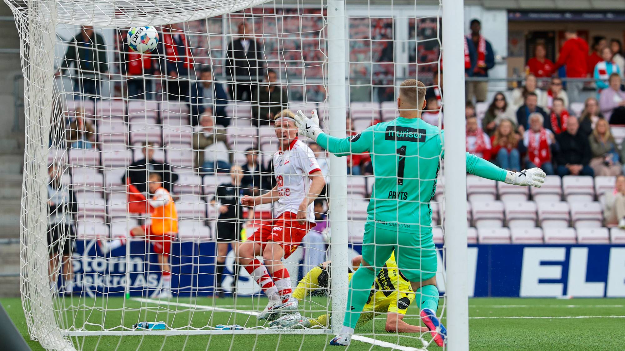 En fotballspiller i rødt og hvitt scorer et mål. Målvakten i grønn drakt forsøker å stoppe ballen. En annen målvakt ligger på bakken. Tilskuere ser på fra tribunen i bakgrunnen. (Bildebeskrivelsen er laget av en KI-tjeneste)