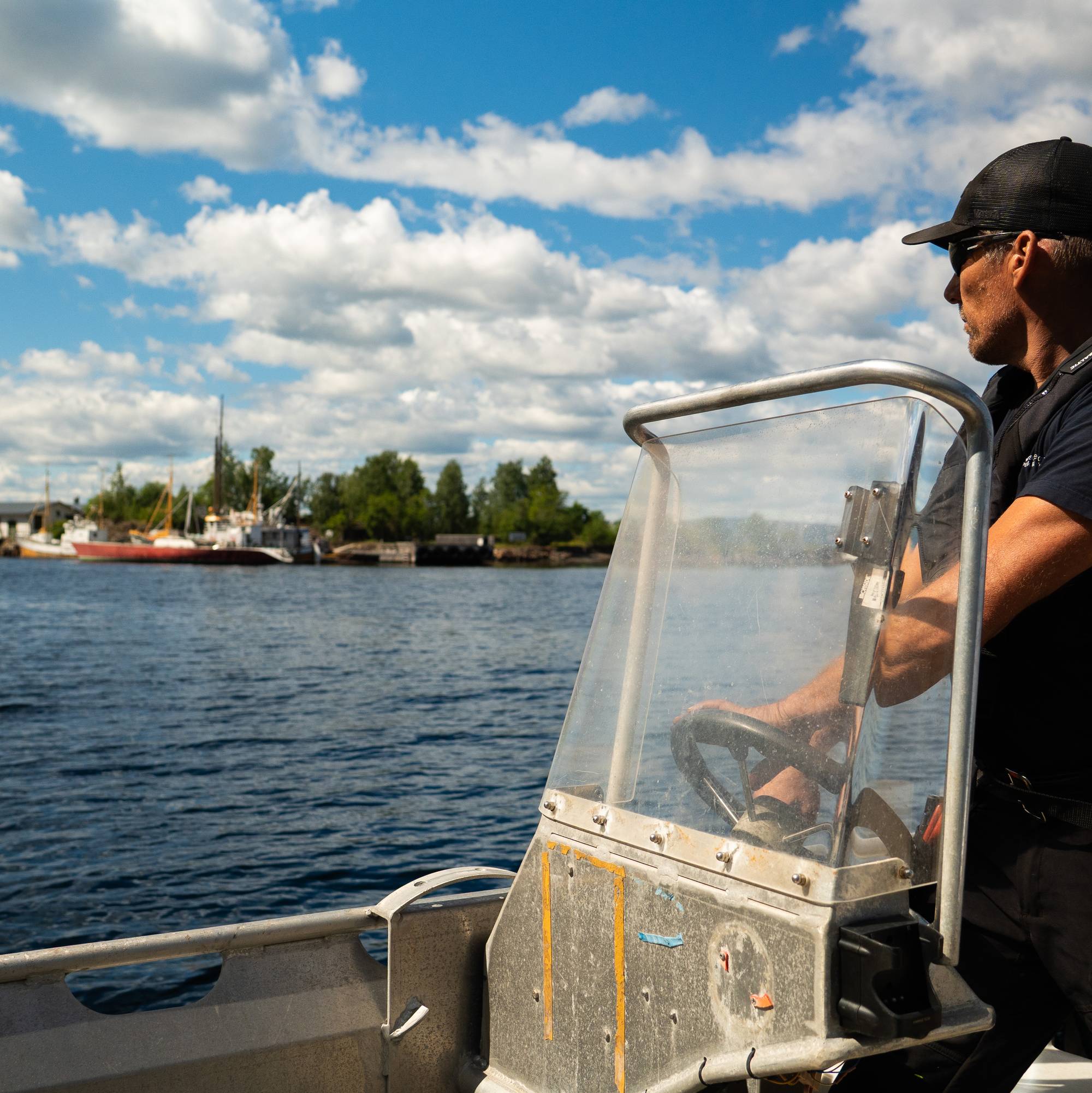 Mann med skyggelue i skjærgårdsbåt i stål ser mot øyer hvor båter har lagt til
