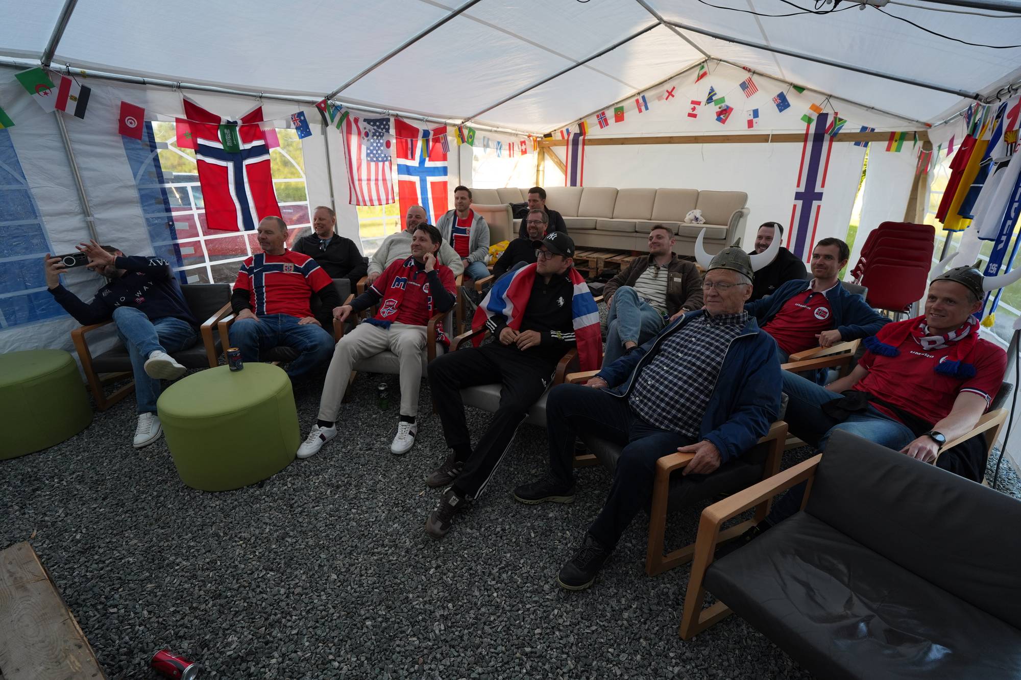 En gruppe menn sitter under et telt og ser på noe. De er iført supporterklær og det henger nasjonalflagg på teltveggene. Noen av mennene har på seg vikinghjelmer. De sitter i stoler på et grusdekke. (Bildebeskrivelsen er laget av en KI-tjeneste)