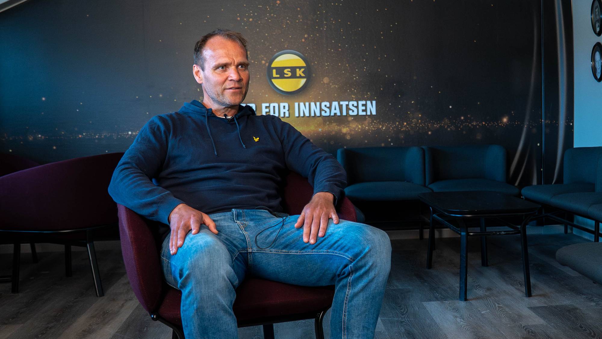 En mann sitter på en mørk sofa foran en vegg med ovale portrettbilder. Bildene viser ansiktene til flere personer i rammer. Bakgrunnen er en lyseblå vegg.