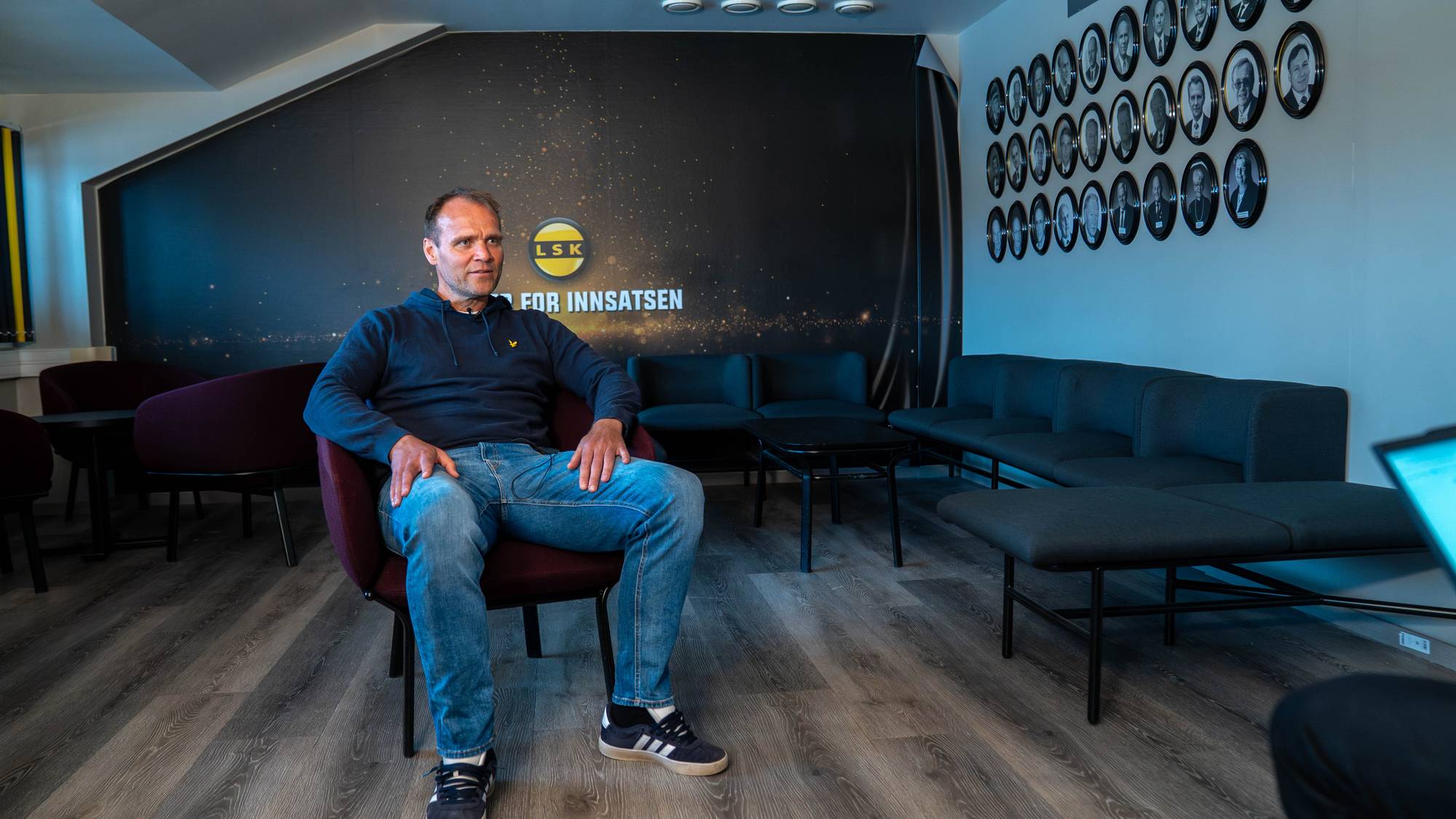 En mann sitter på en mørk sofa foran en vegg med ovale portrettbilder. Bildene viser ansiktene til flere personer i rammer. Bakgrunnen er en lyseblå vegg.