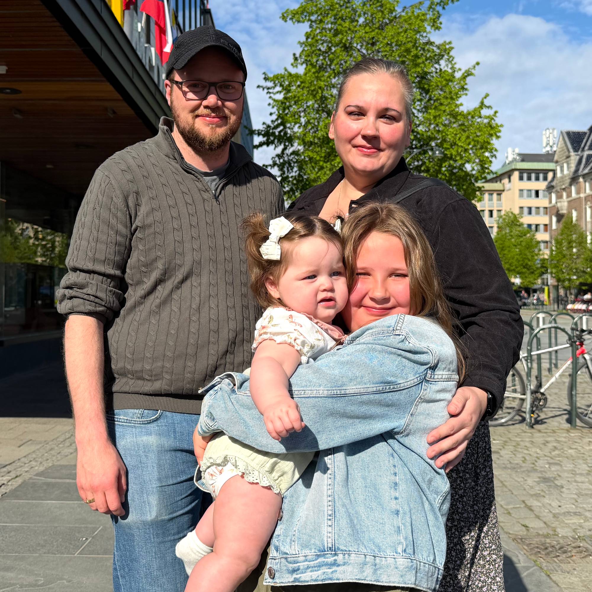 .En familie på fire, far, mor, jente på ni år og jente på åtte måneder, står oppstilt. Foreldrene står bak jentene, den eldste jenta holder den yngste på armen . De smiler til kamera. 