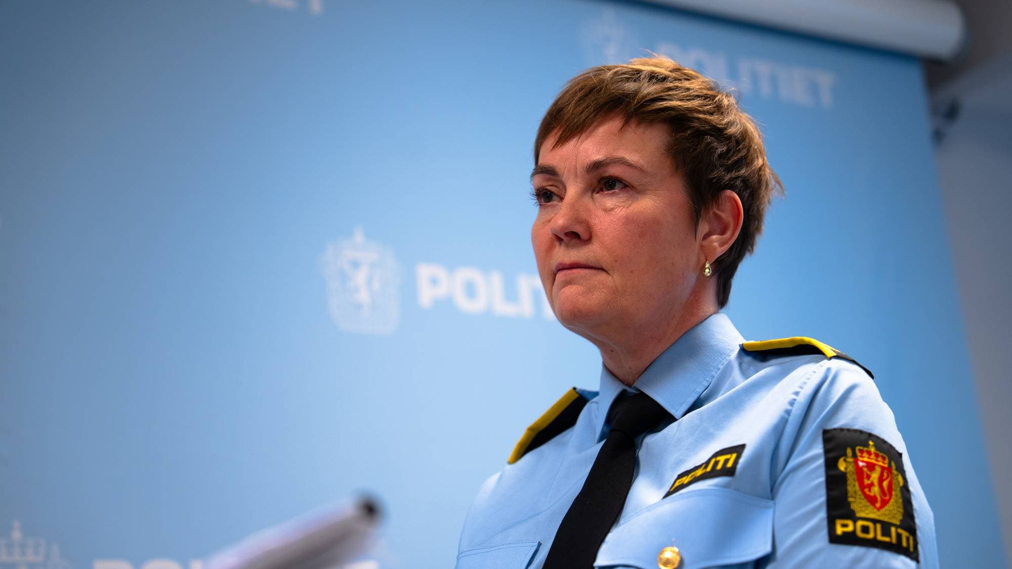 En politibetjent står foran en lyseblå vegg med politiets logo. Hun er iført en uniform med detaljer i svart og gult. Hennes hender er synlige, og hun holder en notisblokk. Det ser ut som hun deltar i en pressekonferanse. (Bildebeskrivelsen er laget av en KI-tjeneste)