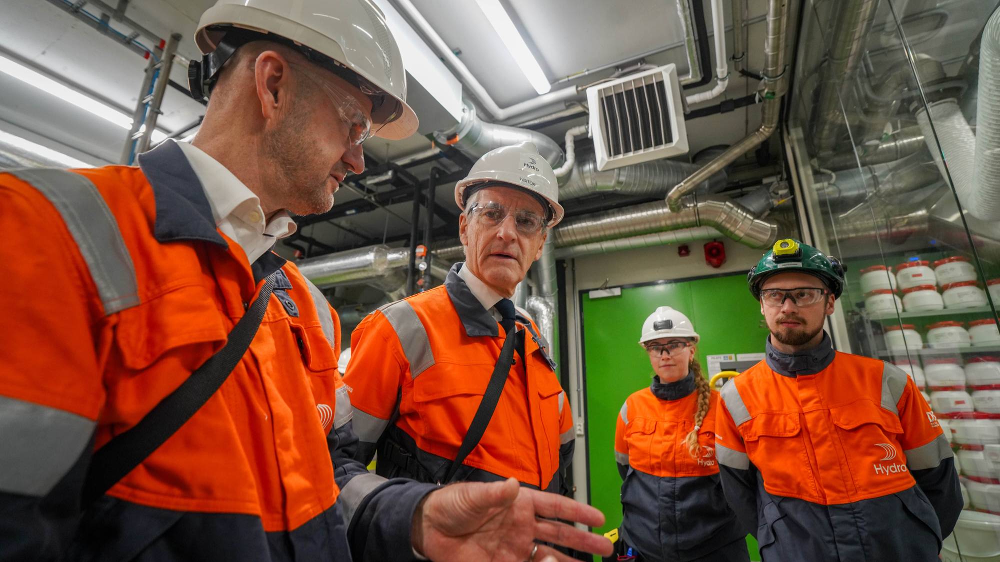 Jonas Gahr Støre på Hydro-besøk sammen med ansatte i verneutstyr i et industrilokale. (Bildebeskrivelsen er laget av en KI-tjeneste)