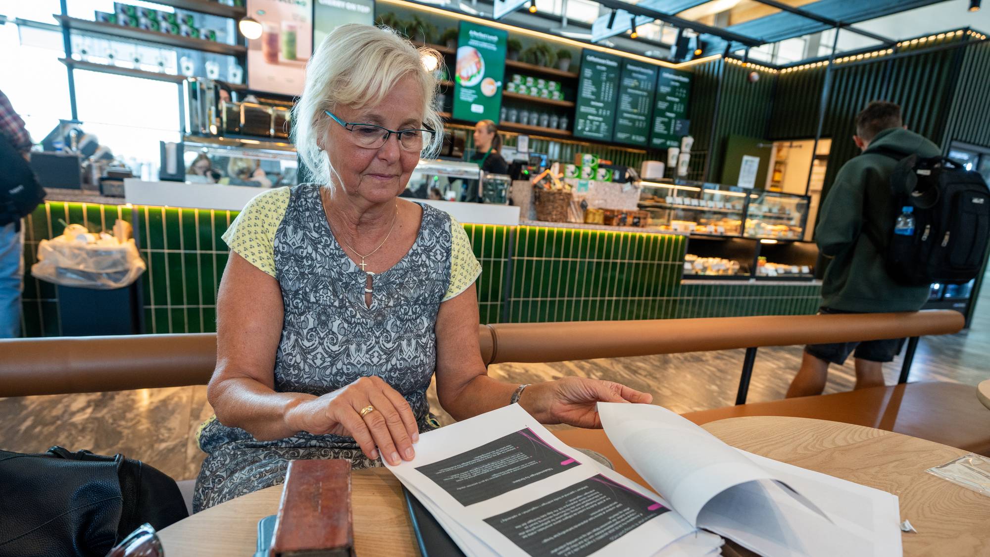 Ei kvinne sitt ved eit bord på ein kafe. Ho har på seg ein grå og gul kjole, ho har lyst hår og briller. På bordet ligg fleire ark som ho blar i. I forgrunnen ser ein mobilen og solbrillene hennar. I bakgrunnen kan ein sjå disken på kafeen. 