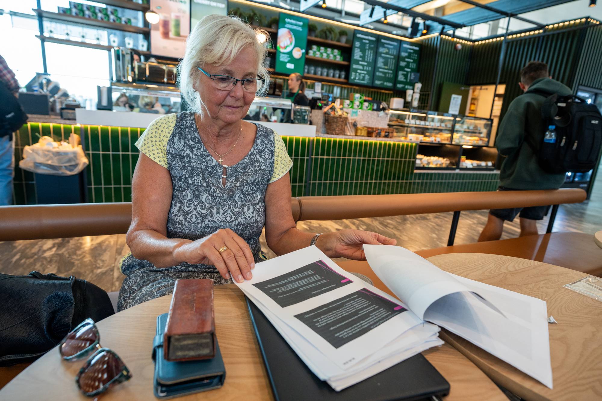 Ei kvinne sitt ved eit bord på ein kafe. Ho har på seg ein grå og gul kjole, ho har lyst hår og briller. På bordet ligg fleire ark som ho blar i. I forgrunnen ser ein mobilen og solbrillene hennar. I bakgrunnen kan ein sjå disken på kafeen. 