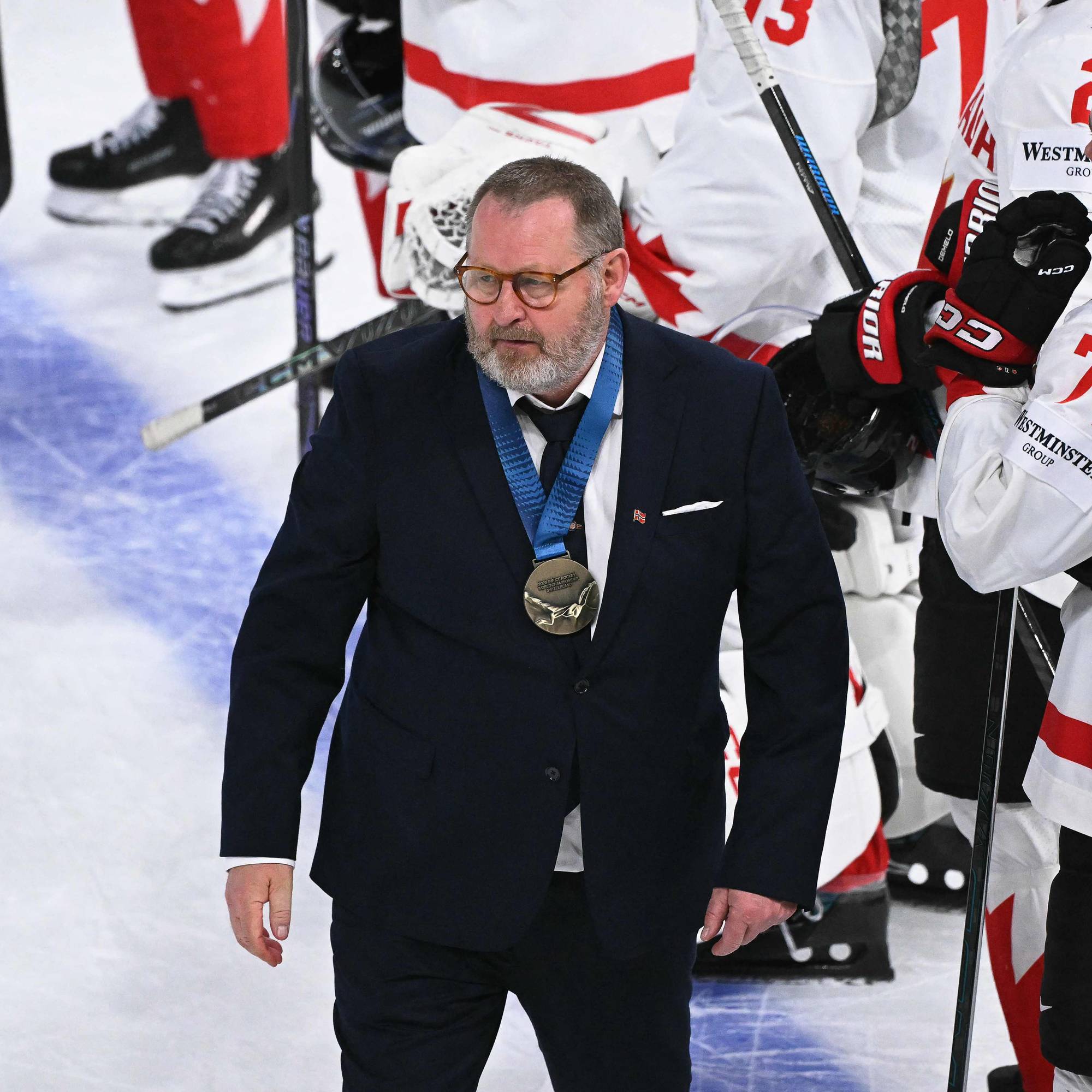 En mann med dress og medalje går på isen. Han er omgitt av ishockeyspillere i hvite drakter med røde detaljer. Flere spillere har nummer synlig på draktene. En av spillerne har navnet "CELEBRINI" på drakten. (Bildebeskrivelsen er laget av en KI-tjeneste)