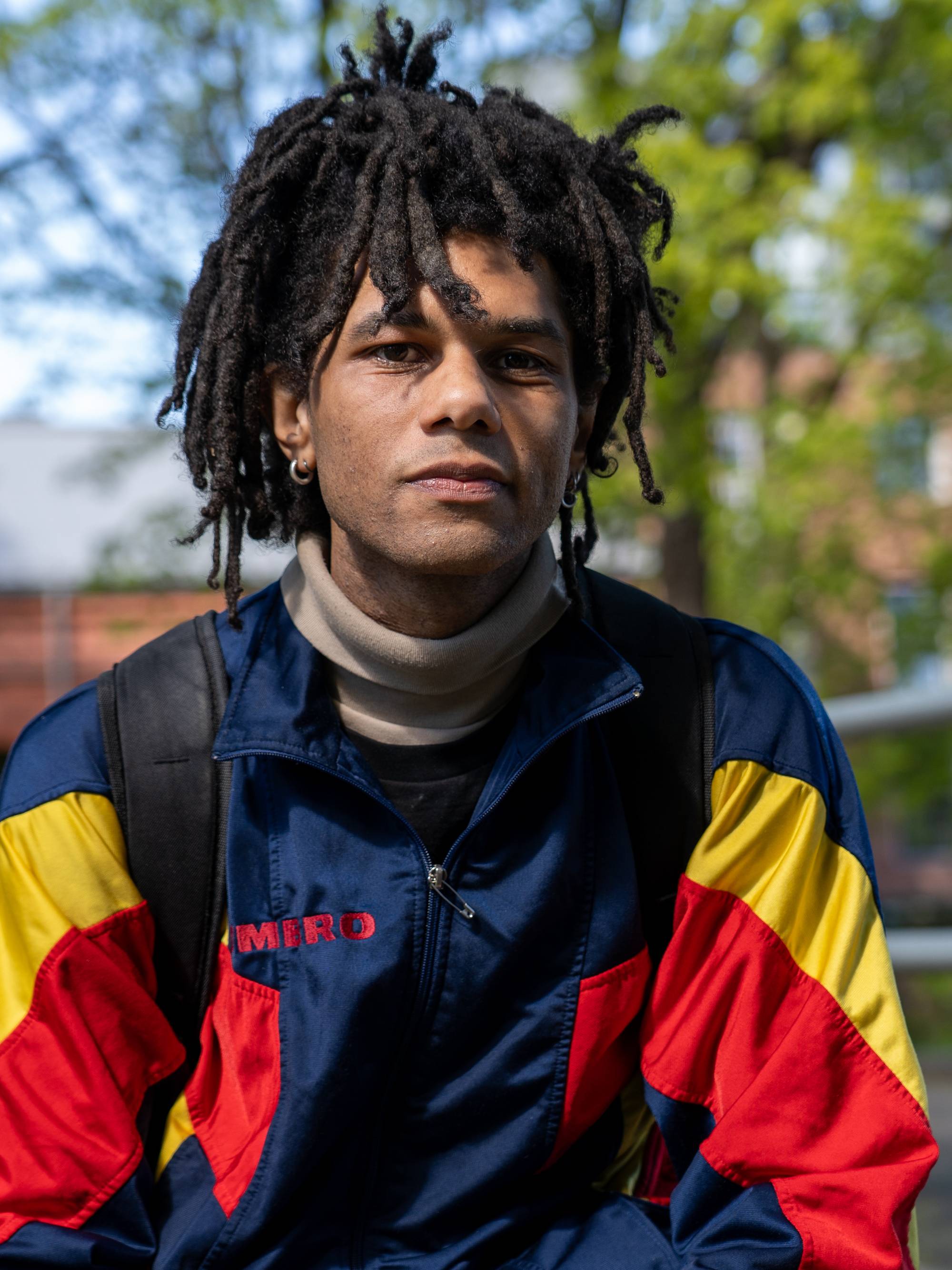 Et bilde av en mann med dreadlocks som sitter utendørs. Han har på seg en blå, rød og gul jakke. I bakgrunnen er det trær og en bygning. (Bildebeskrivelsen er laget av en KI-tjeneste)
