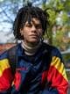 Audun Torsdalen / NRK Et bilde av en mann med dreadlocks som sitter utendørs. Han har på seg en blå, rød og gul jakke. I bakgrunnen er det trær og en bygning. (Bildebeskrivelsen er laget av en KI-tjeneste)