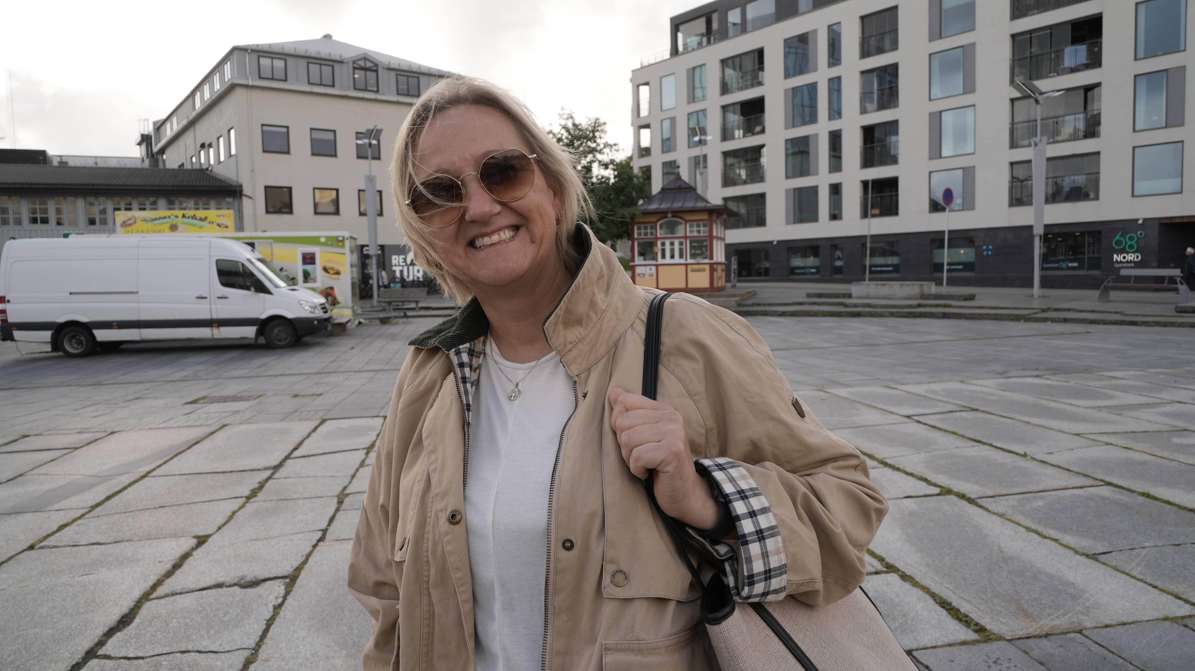 En dame med solbriller og kort lyst hår smiler til kamera. Hun har hvit t-skjorte og beige jakke og veske. I bakgrunnen synes en varebil og flere hvite bygninger.