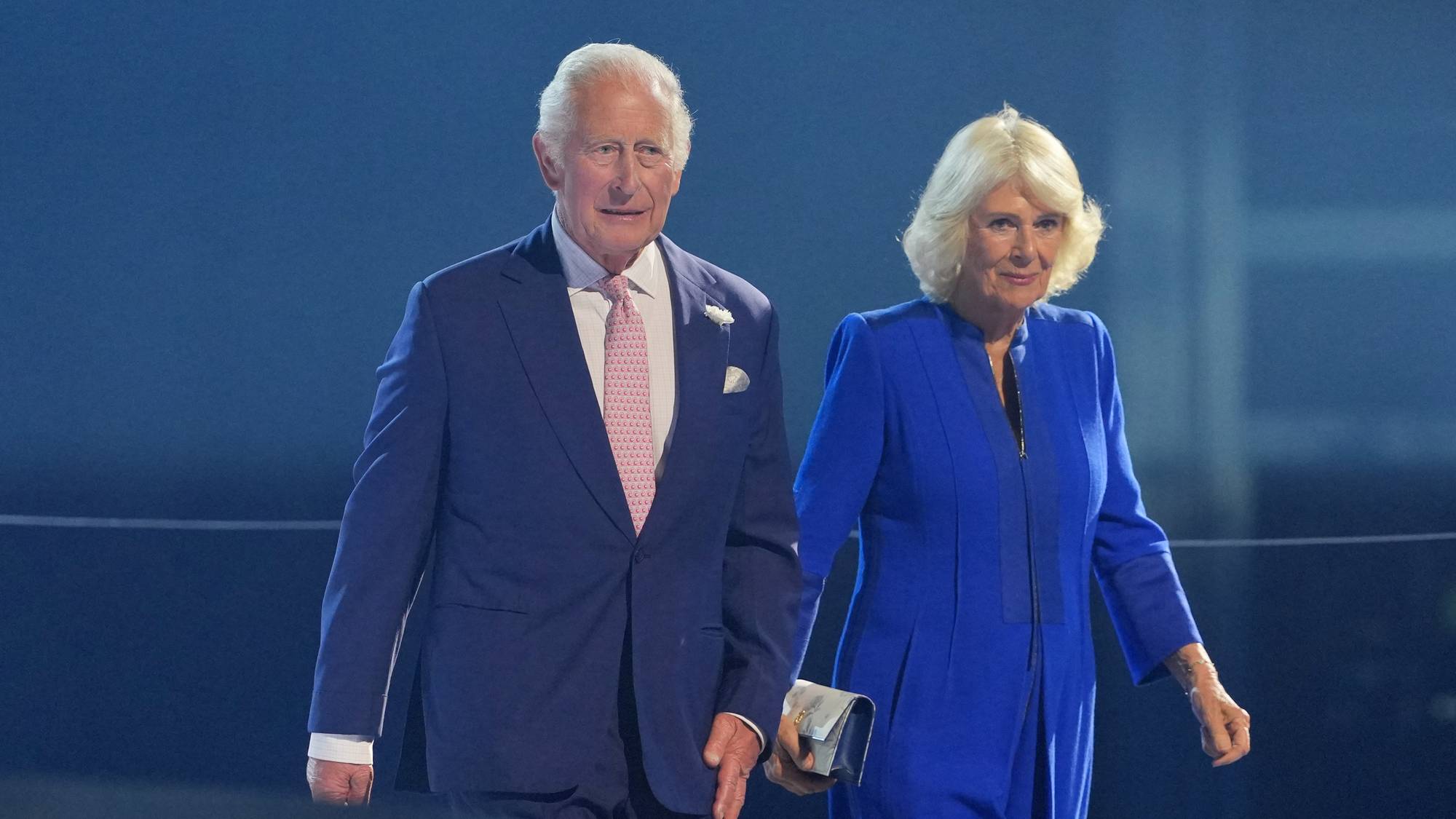 Kong Charles og dronning Camilla står ved siden av hverandre mot en blå bakgrunn. 
