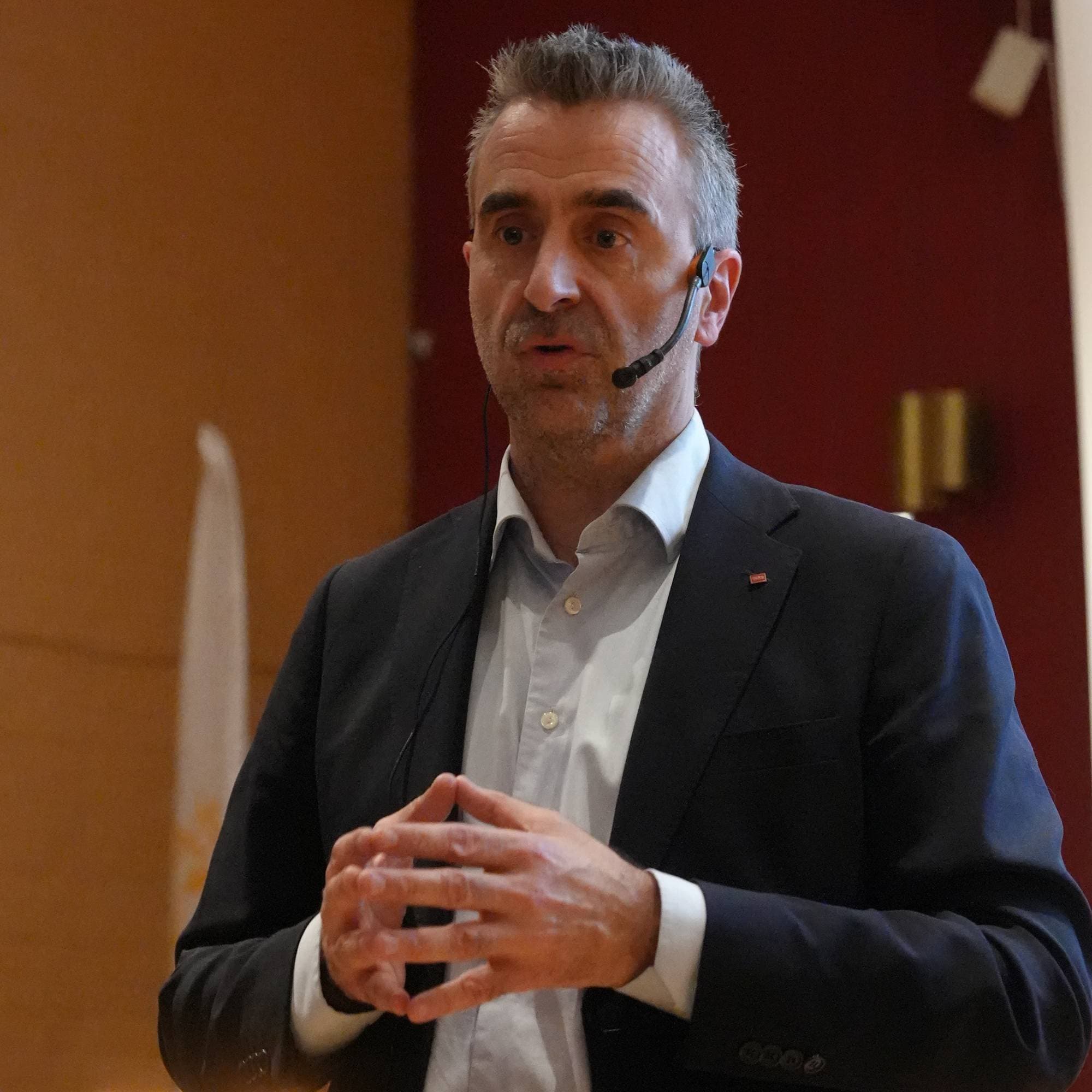 En mann i mørk dress og hvit skjorte står og foredrar, han har en mikrofon på siden av ansiktet