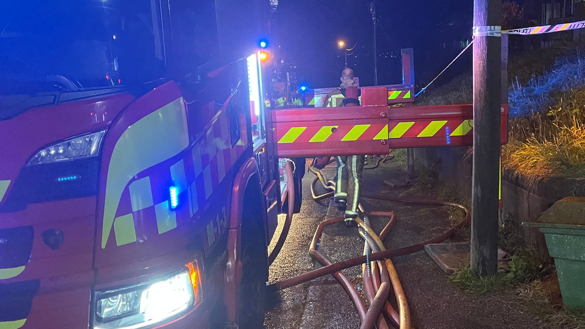Brannvesenet på stedet ved en rød brannbil med slanger utlagt på bakken i mørket. (Bildebeskrivelsen er laget av en KI-tjeneste)