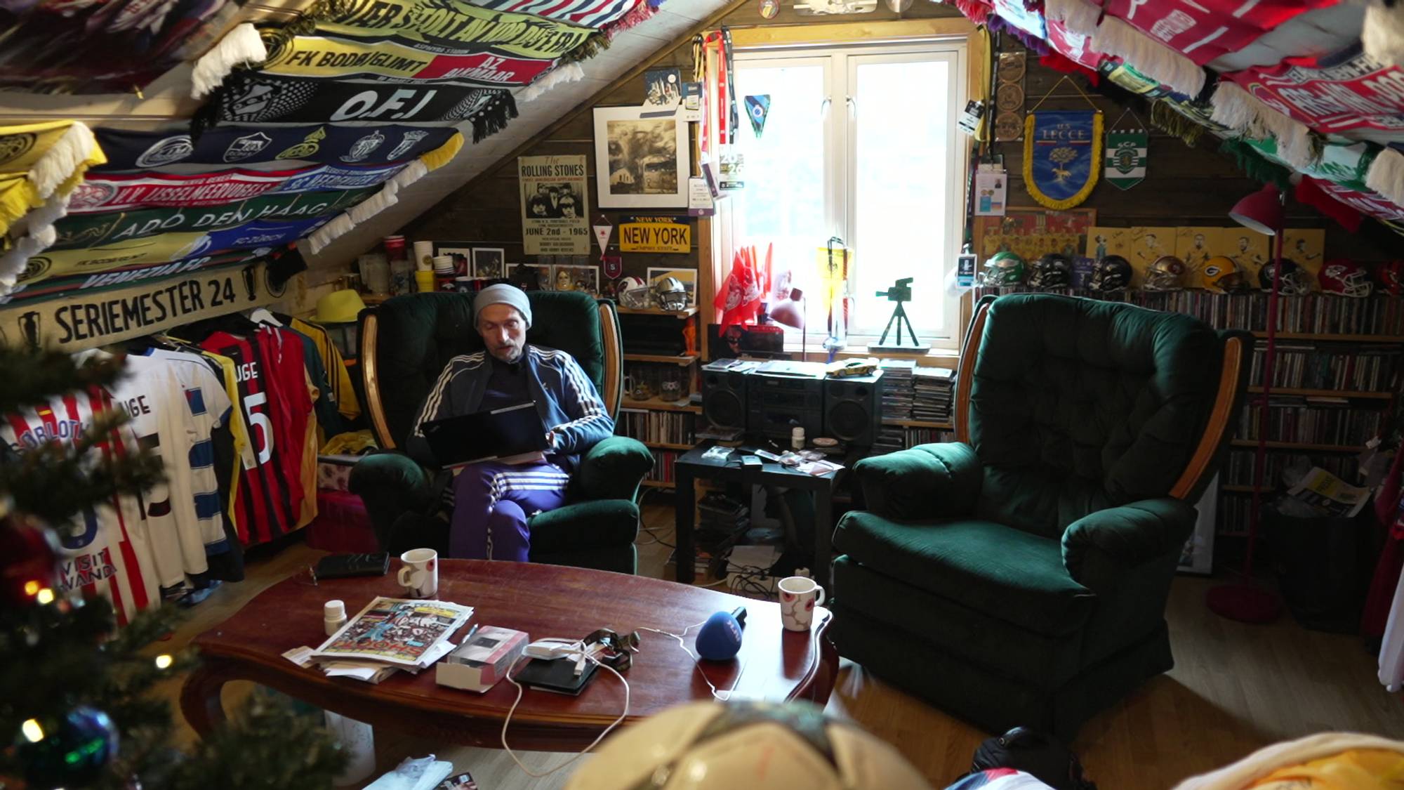 En mann sitter i en grønn lenestol og bruker en datamaskin. Rommet er dekorert med fotballdrakter og supporter-skjerf. Flere hyller er fylt med bøker og samleobjekter. Et vindu er synlig bak mannen, med et tre utenfor. (Bildebeskrivelsen er laget av en KI-tjeneste)
