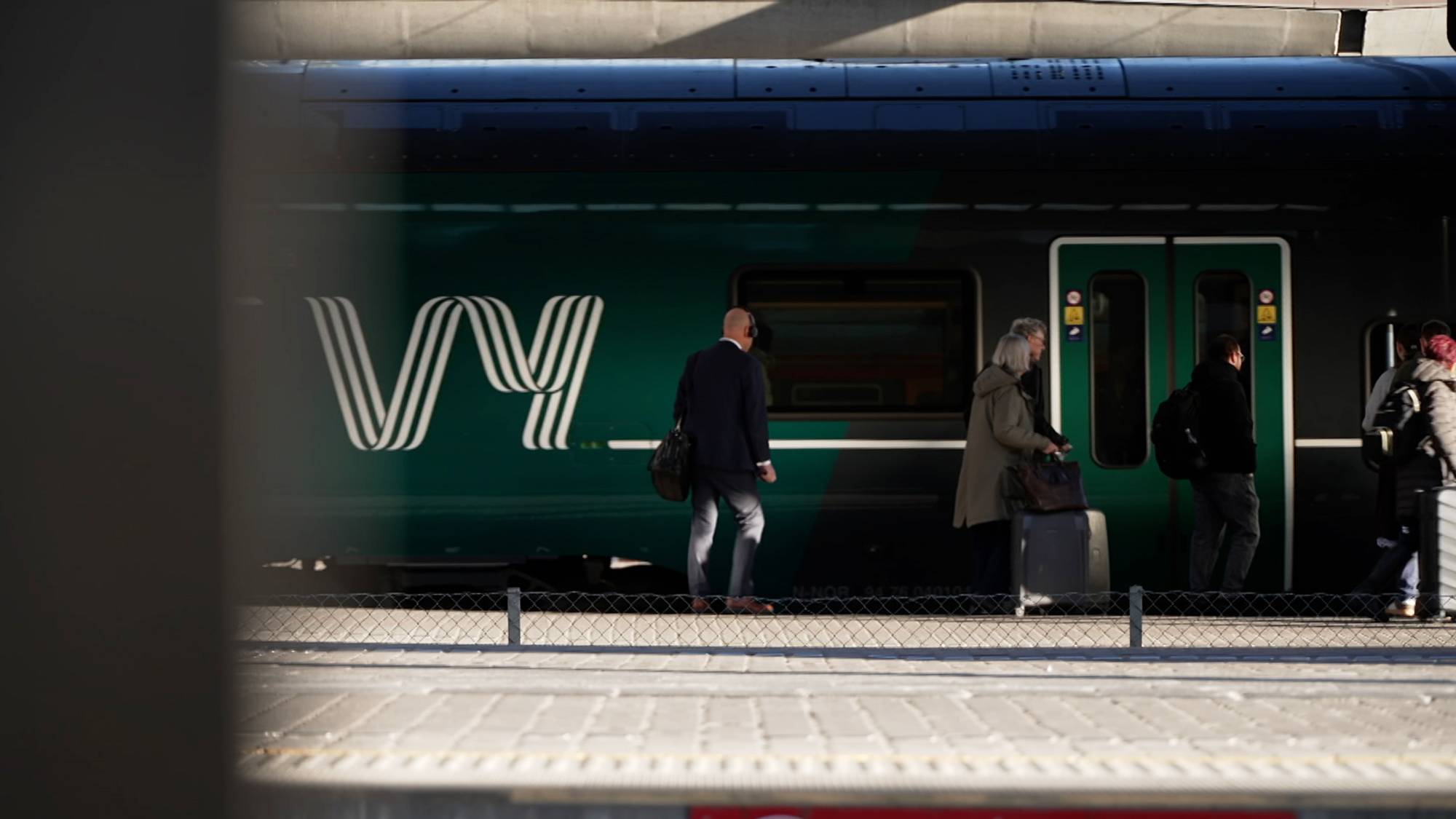 En gruppe reisende venter ved en togstasjon, med en tog vogn synlig i bakgrunnen. Flere personer bærer bagasje og er i ferd med å gå mot toget, mens en kvinne med lyst hår står med en koffert. Den synlige togvognen har en grønn farge med hvite detaljer og er merket med logoen "vy". Omgivelsene er preget av en moderne stasjon med fliser på bakken. (Bildebeskrivelsen er laget av en KI-tjeneste)