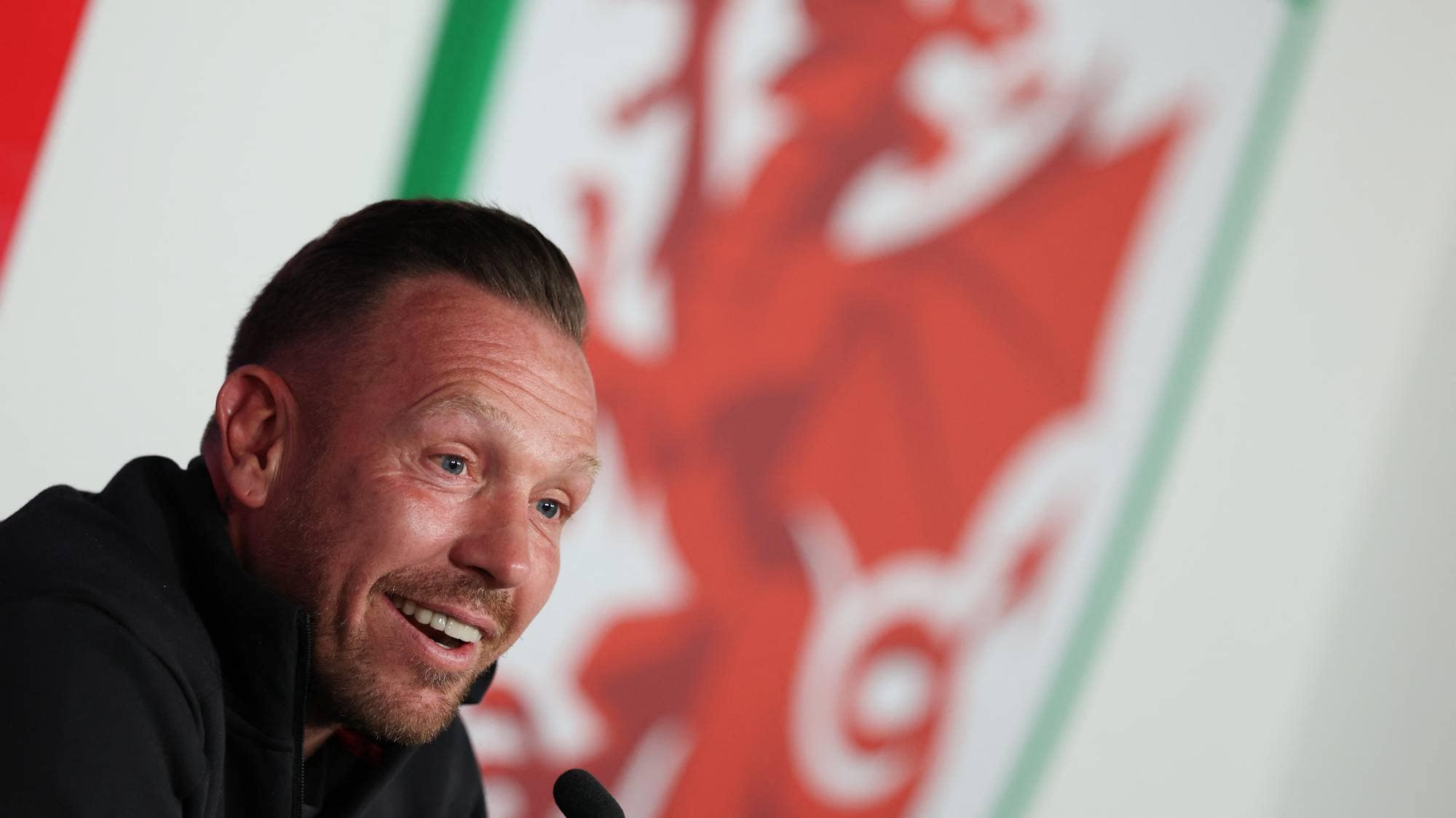 Craig Bellamy smiler foran det walisiske fotballforbundets emblem på en pressekonferanse. (Bildebeskrivelsen er laget av en KI-tjeneste)