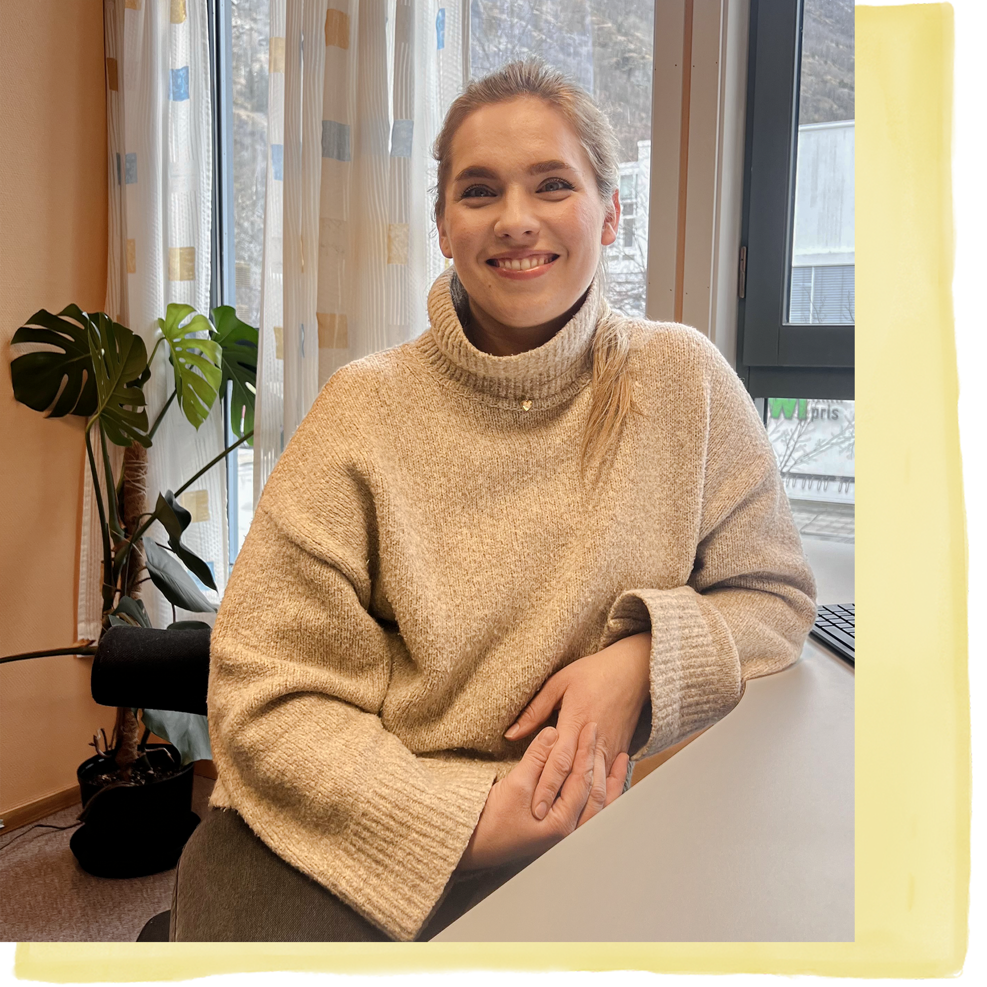 Kvinne sitter ved pulten. Hun ser i kamera og smiler.