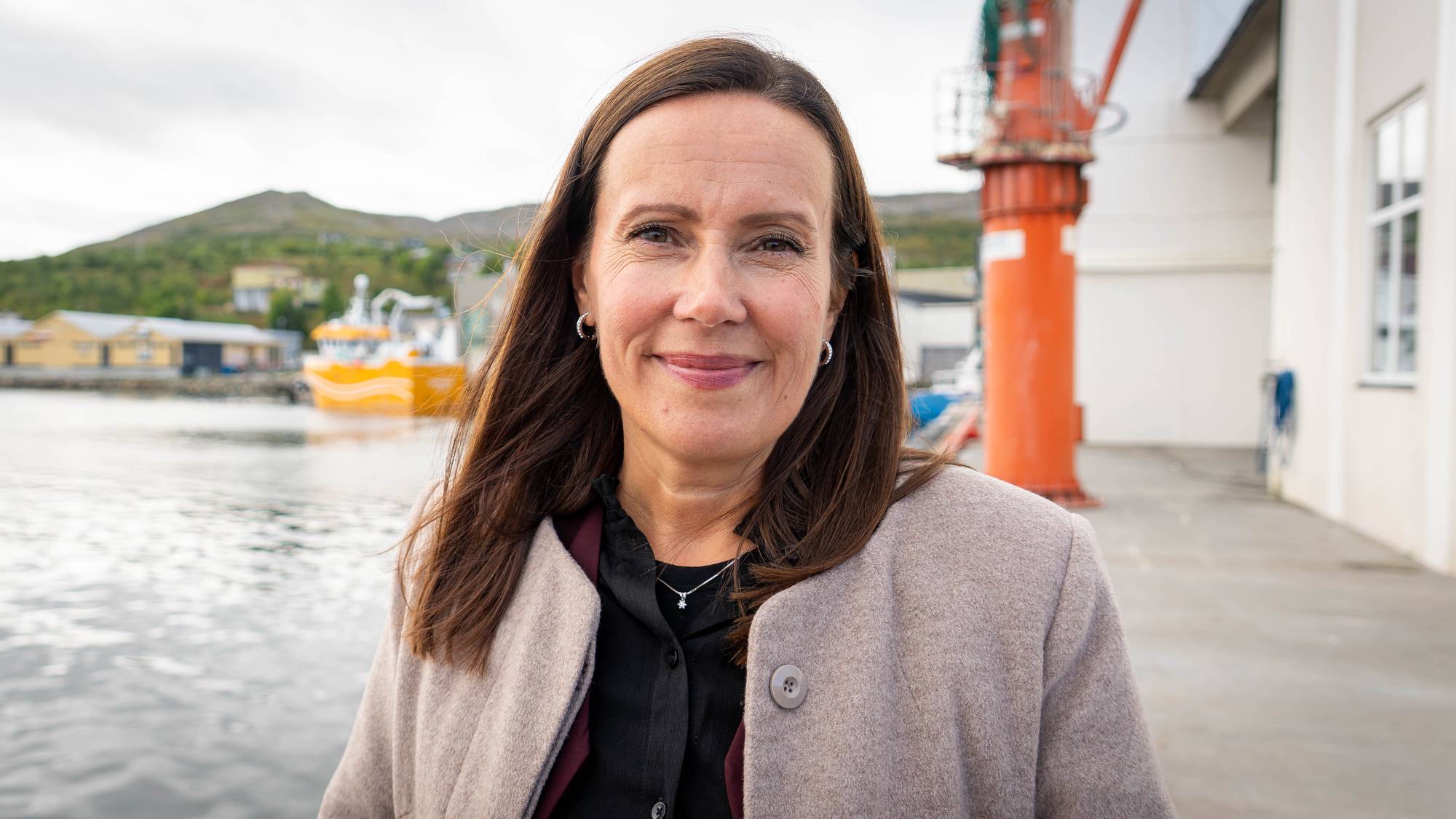 Kvinne med mørkt hår fotografert utendørs ved en havn med fiskebåt i bakgrunnen. (Bildebeskrivelsen er laget av en KI-tjeneste)
