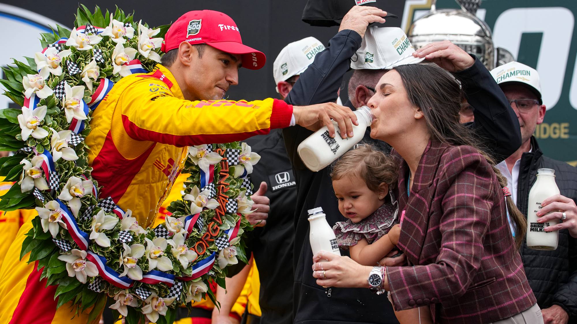 En fører i gul racerdrakt mottar en seierskrans med blomster og bånd. En kvinne holder en baby og drikker fra en melkeflaske. Andre personer er bak dem. (Bildebeskrivelsen er laget av en KI-tjeneste)