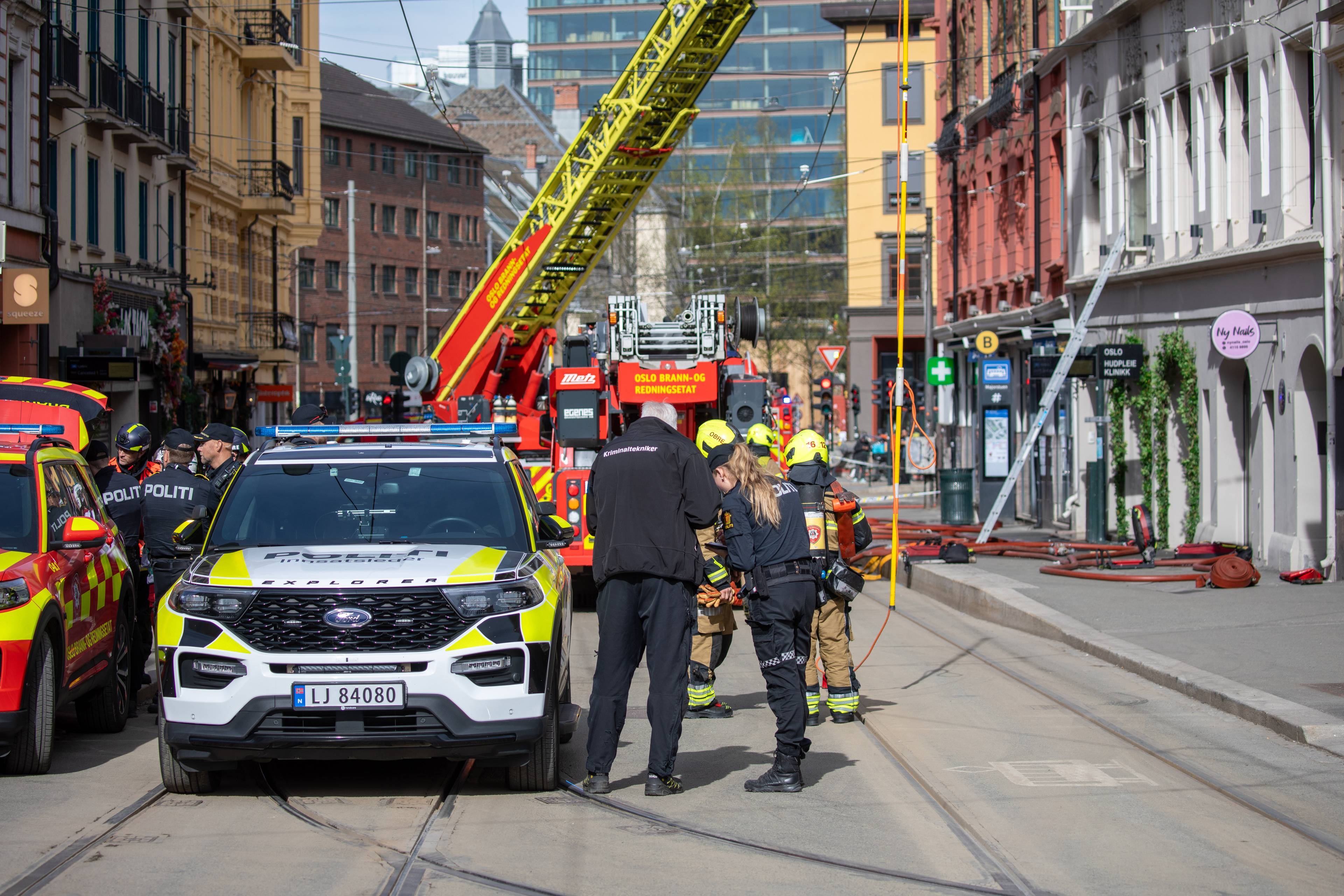 Politi og brannvesen er på stedet. En brannbil med stige er parkert ved siden av en politibil. Flere personer i uniform står samlet ved politibilen. Slanger ligger på bakken i gaten. (Bildebeskrivelsen er laget av en KI-tjeneste)