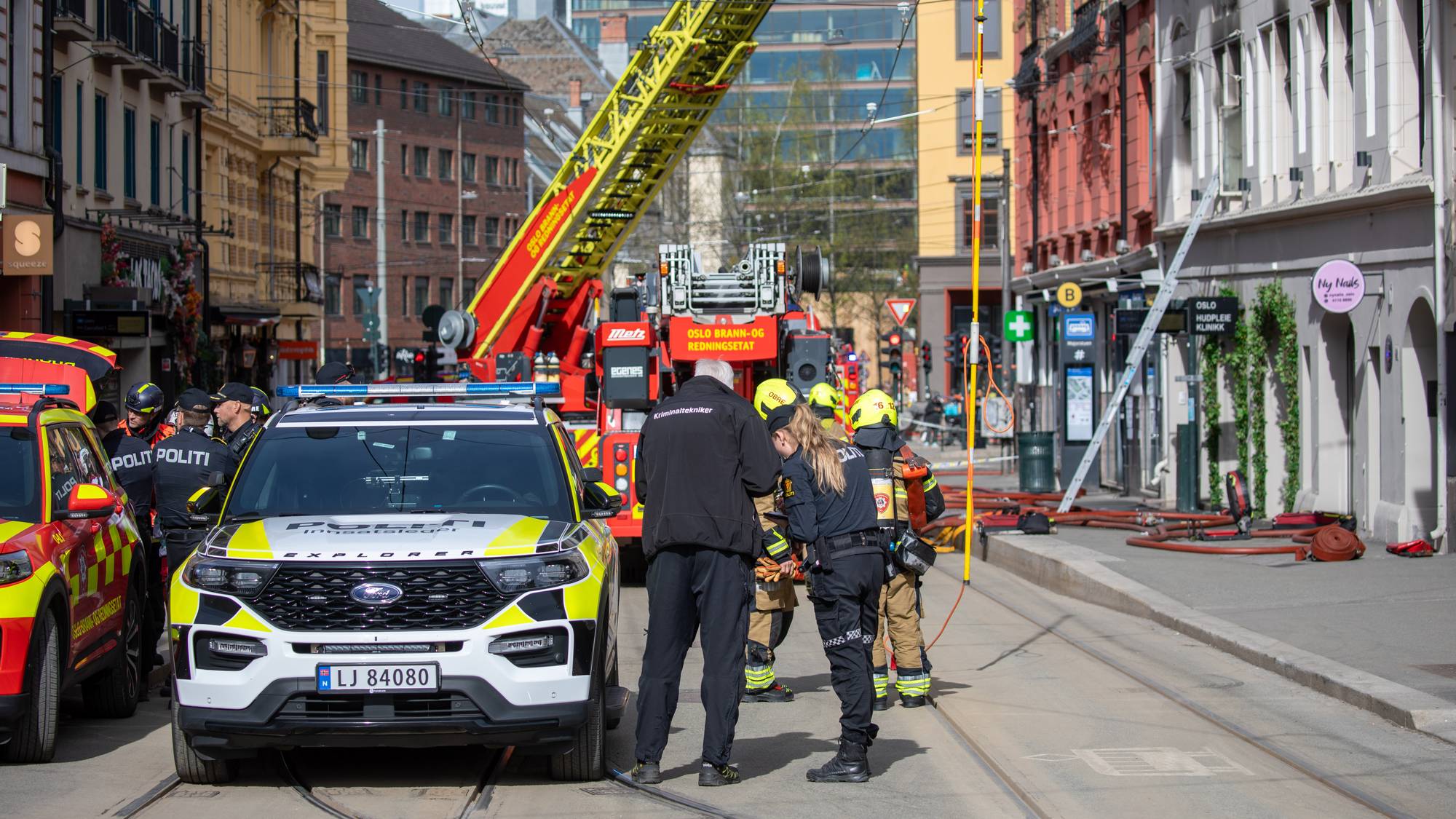 Politi og brannvesen er på stedet. En brannbil med stige er parkert ved siden av en politibil. Flere personer i uniform står samlet ved politibilen. Slanger ligger på bakken i gaten. (Bildebeskrivelsen er laget av en KI-tjeneste)