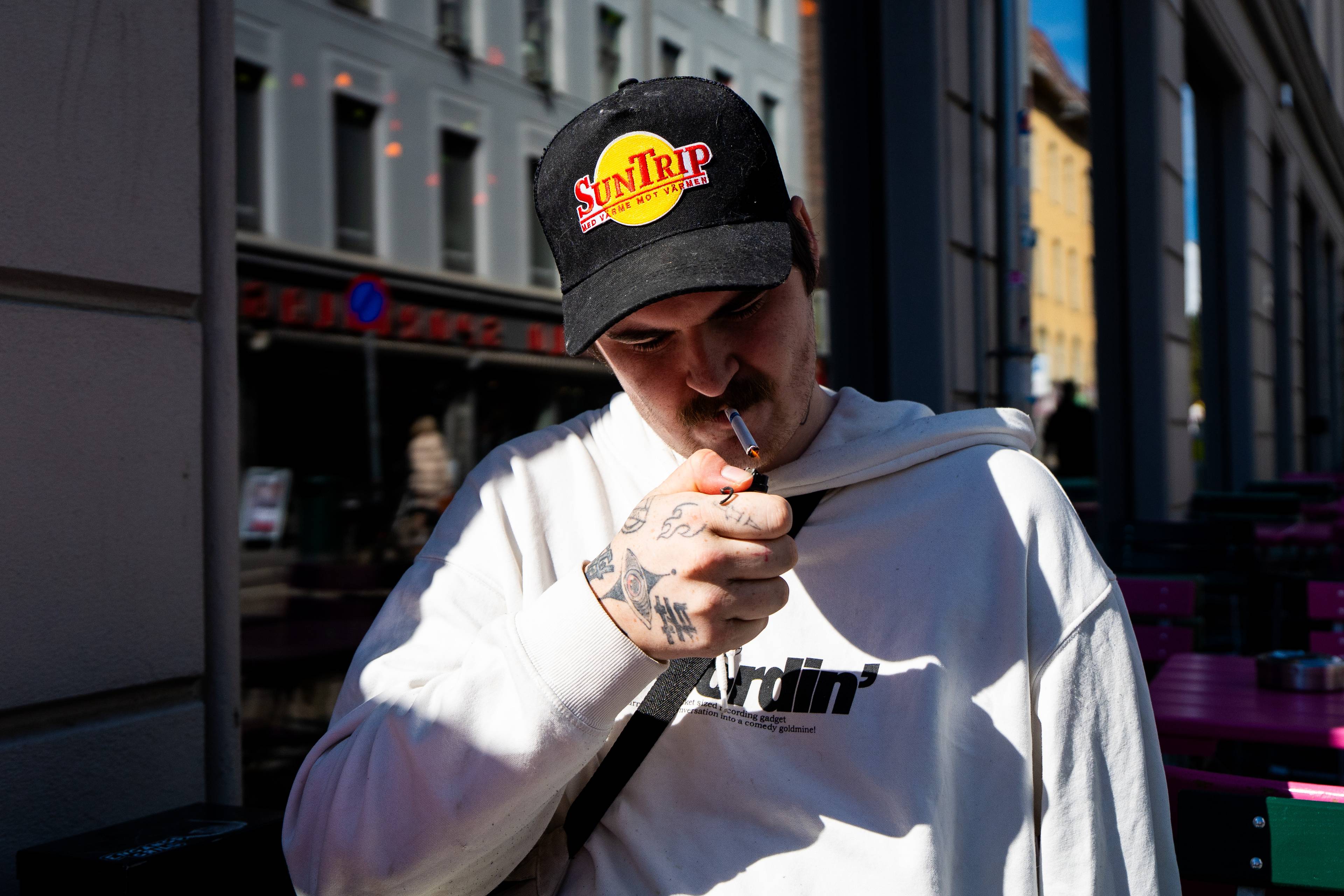 Ein mann i kvit hettegenser og kaps tenner ei sigarett med lighter. Mannen har tatoveringar på hendene. I bakgrunnen sees bygningar og utemøblar. (Bildebeskrivelsen er laget av ei KI-teneste)