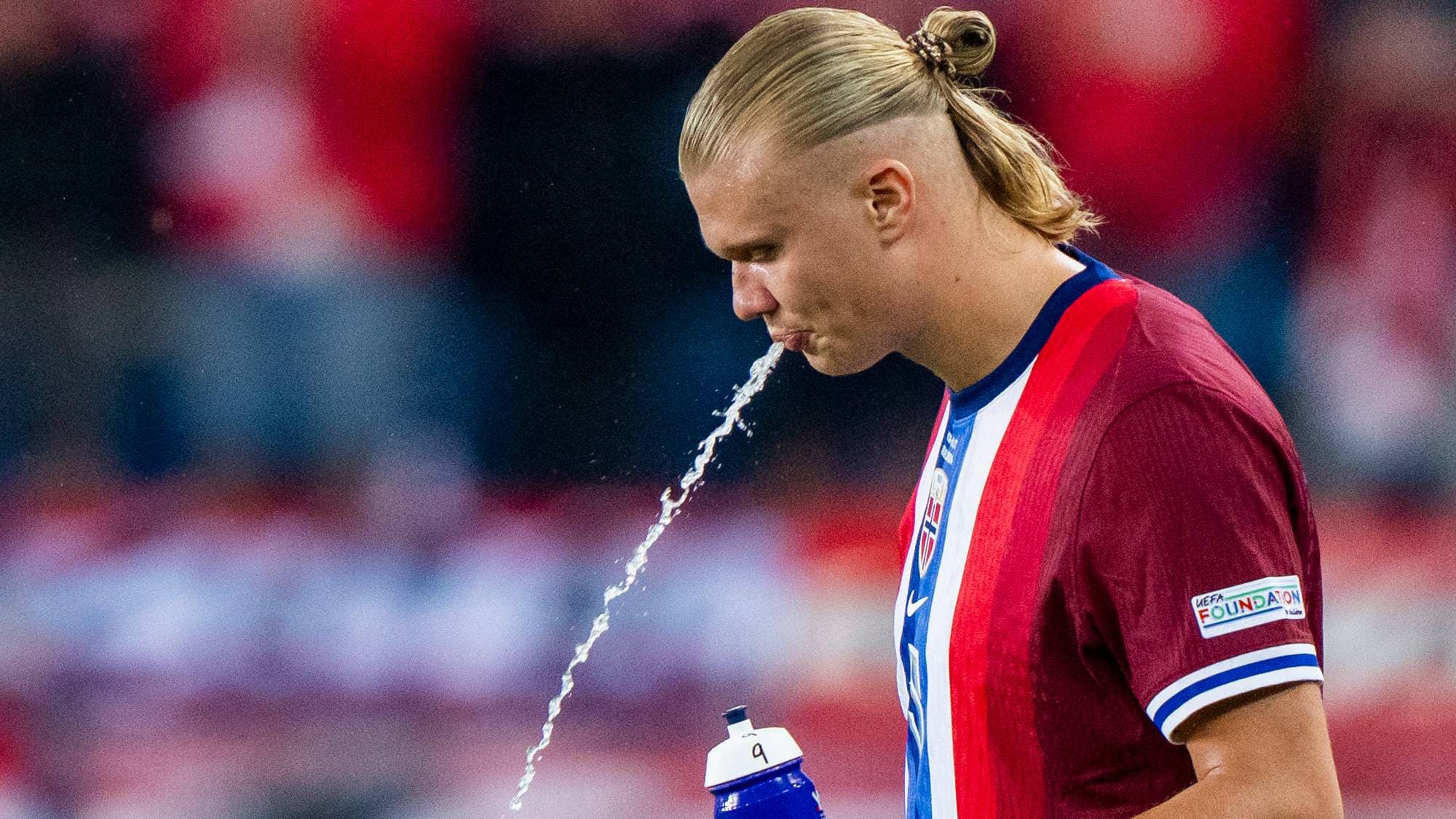 Fotballspilleren Erling Braut Haaland spytter vann. Han holder en blå drikkeflaske i hånden. Bak ham sitter publikum på en tribuna. (Bildebeskrivelsen er laget av en KI-tjeneste)