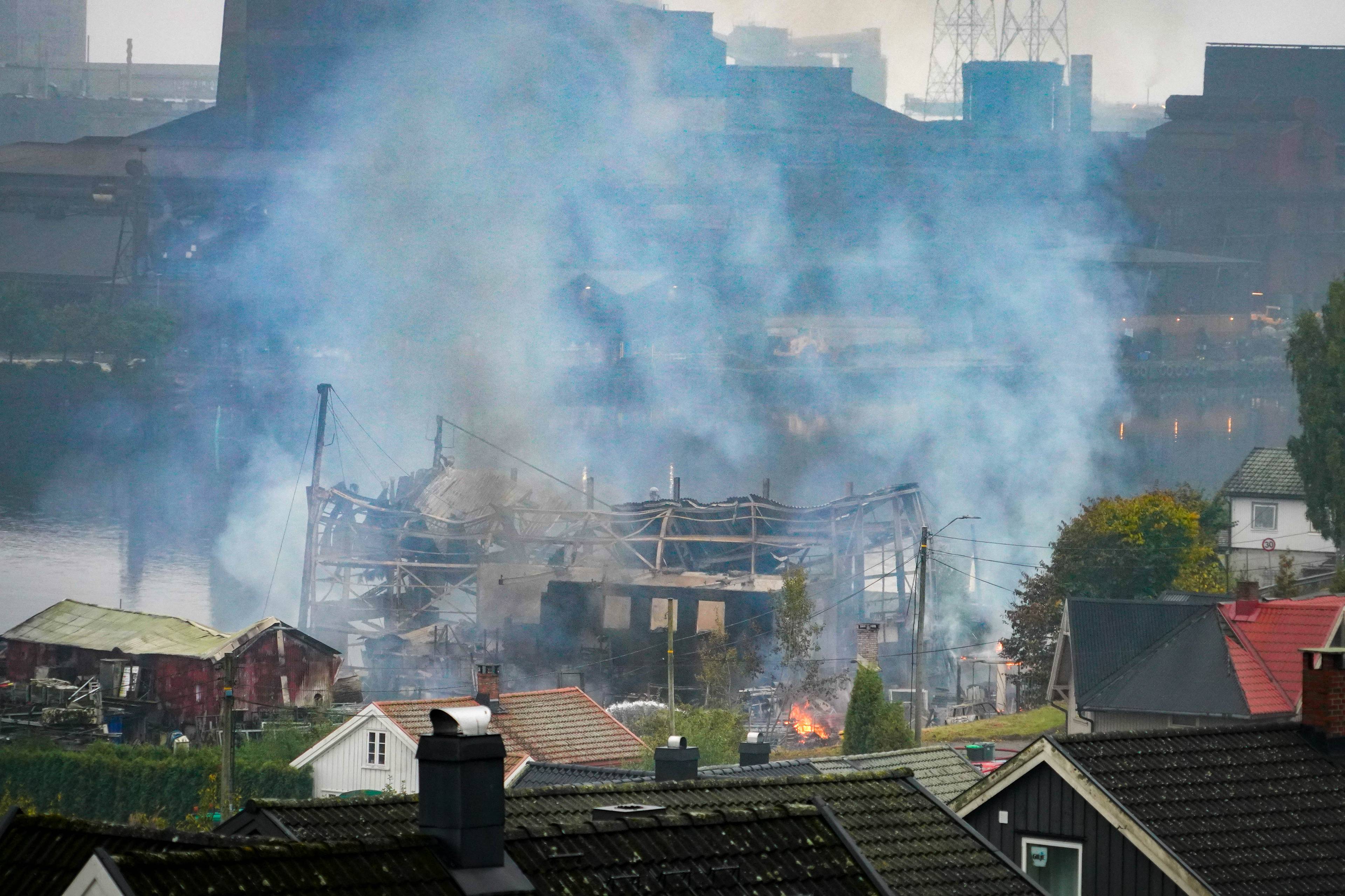 Tykk røyk stiger opp fra en brennende bygning ved vannet. Industribygg er synlige i bakgrunnen. (Bildebeskrivelsen er laget av en KI-tjeneste)